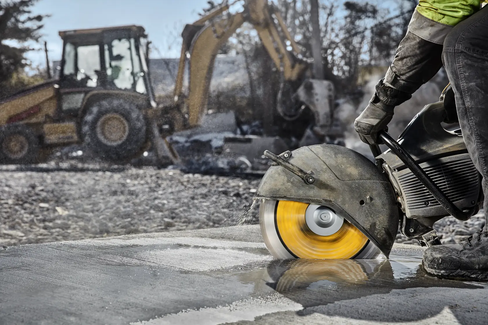A construction worker using a DEWALT concrete saw with a yellow blade to cut a wet concrete surface. In the background, there is construction equipment such as an excavator and a loader.