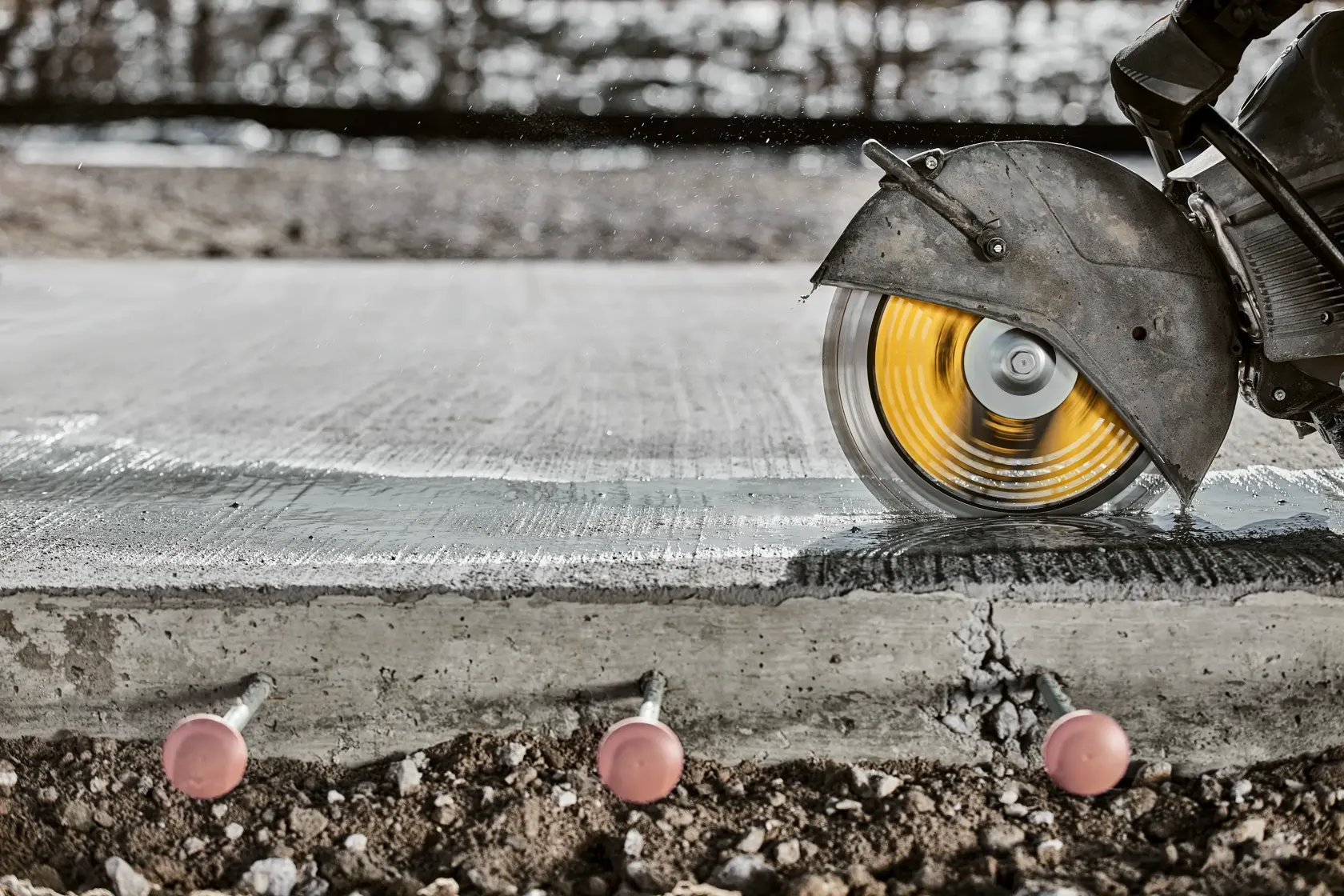 A close-up of a circular saw with a yellow blade cutting through reinforced concrete outdoors. Three metal rods with pink caps are visible in the concrete slab.