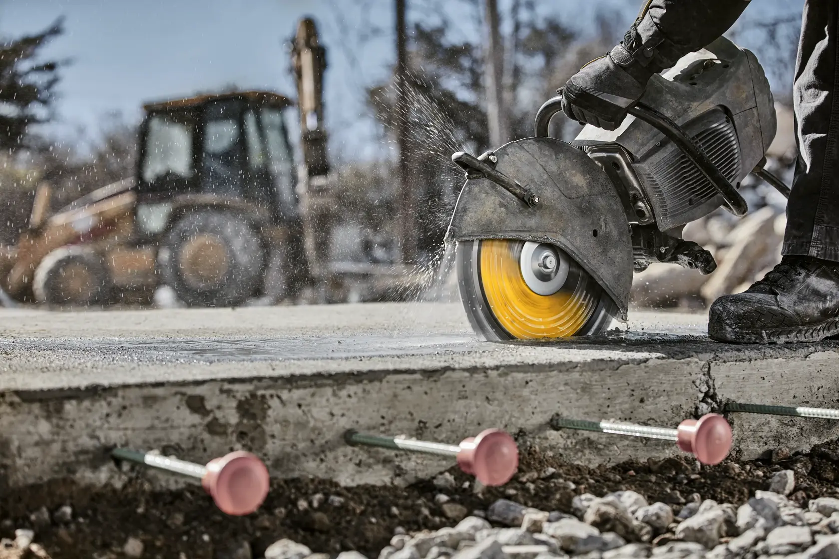 A close-up of a construction worker using a DEWALT power saw with a yellow blade to cut through concrete. Water sprays from the saw during operation. In the background, there is a blurred image of a heavy construction vehicle. Under the concrete, anchor bolts with pink caps are visible.