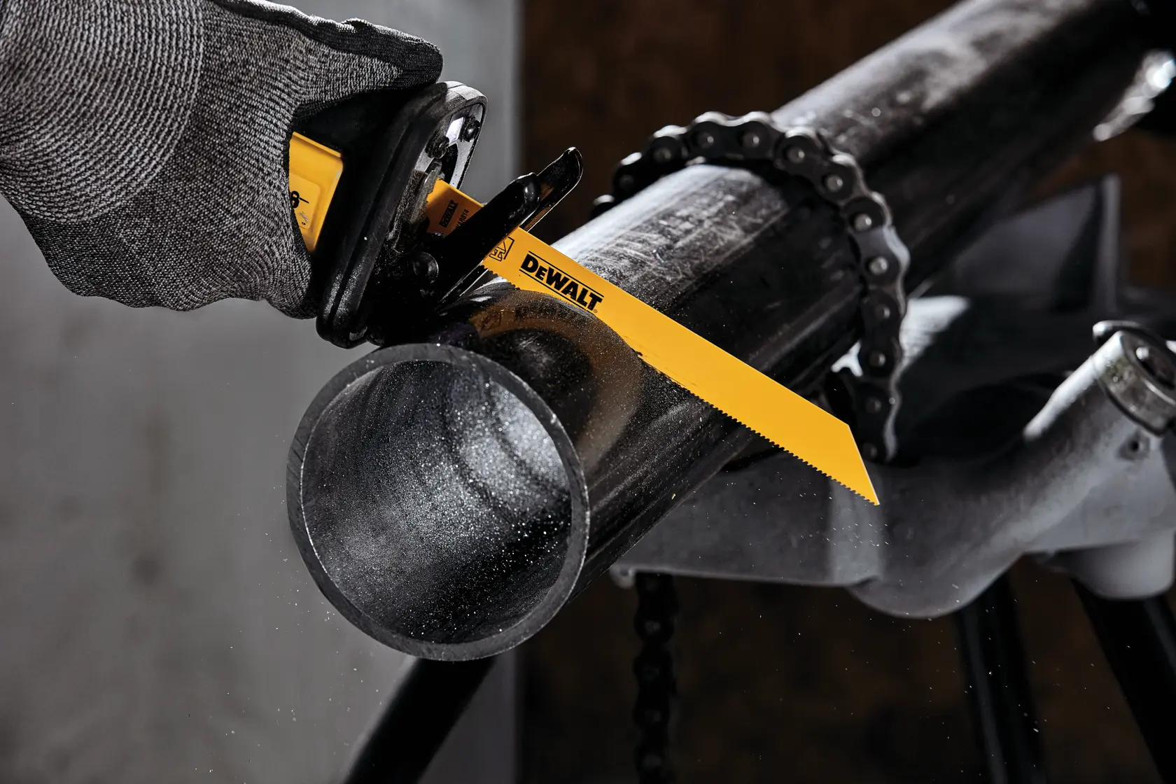 A close-up of a DEWALT reciprocating saw blade cutting through a metal pipe, with a gloved hand holding the power tool and the pipe secured by a chain clamp.