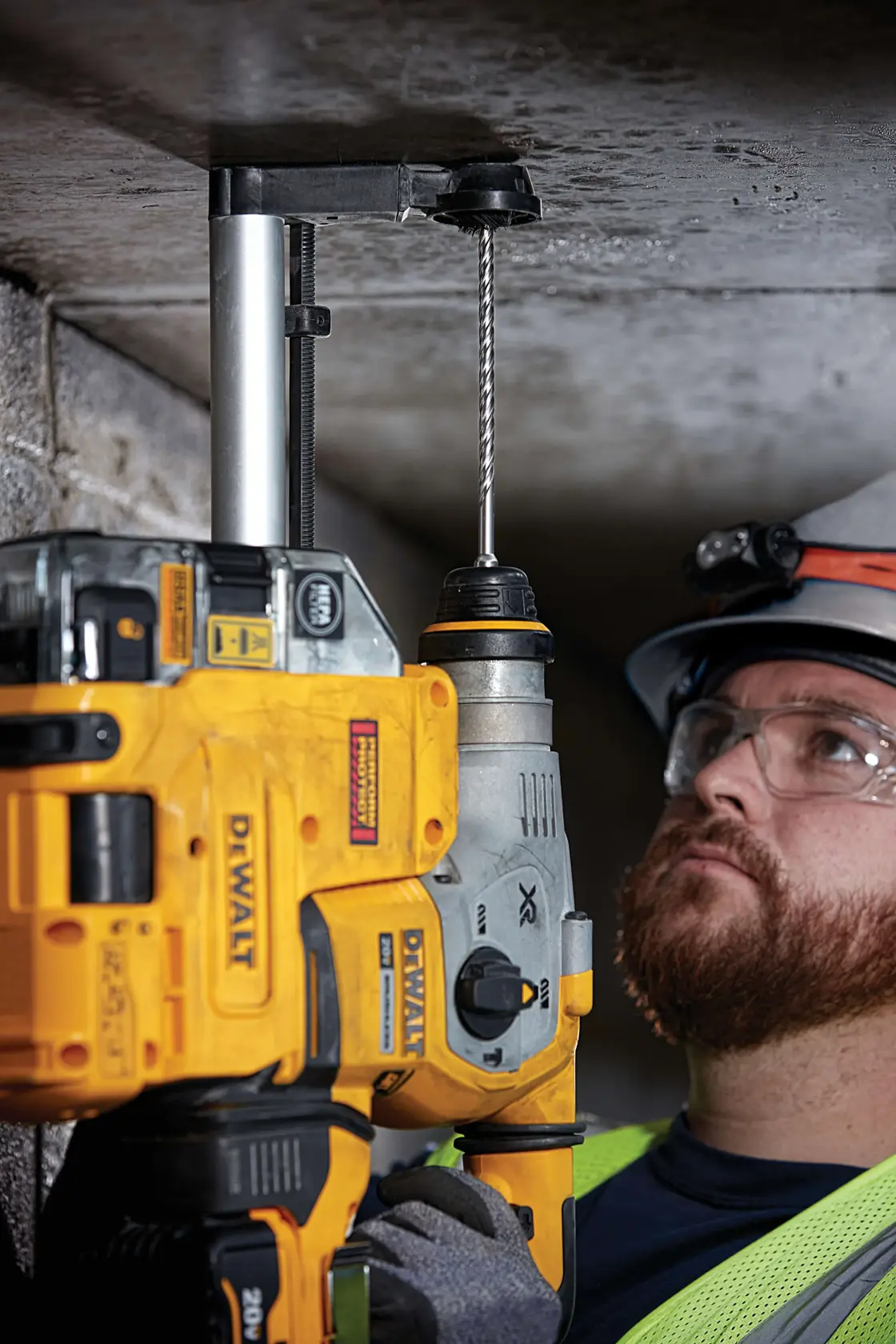 A person using a yellow DEWALT power drill with a drill bit attachment to make a hole in a concrete ceiling. The person is wearing safety equipment including a helmet and hi-vis vest. Their face is intentionally blurred.
