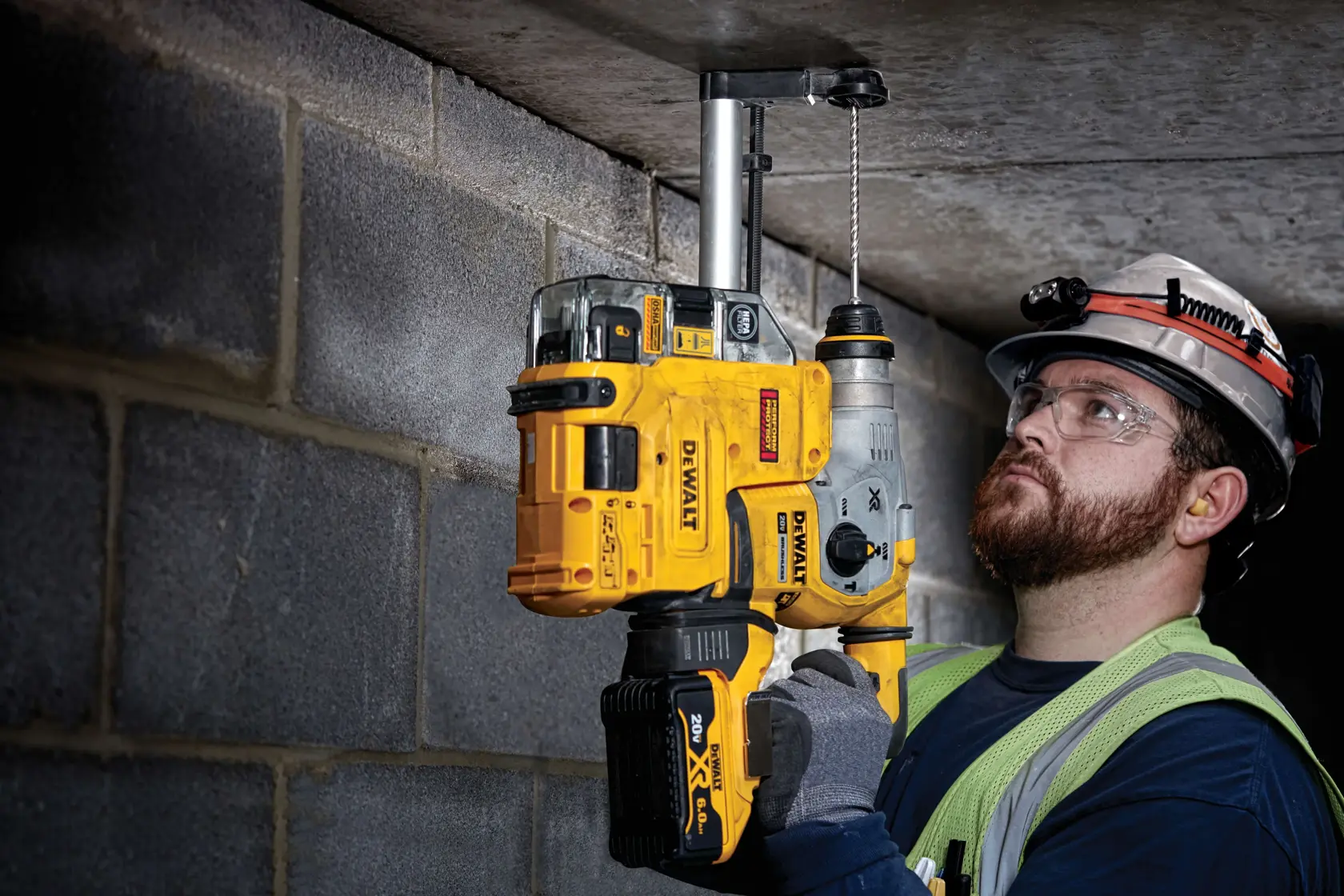 A person wearing construction safety gear uses a DEWALT cordless rotary hammer drill to drill into a concrete ceiling. The drill is yellow and black, and the worker is wearing gloves, a reflective vest, and a helmet with a headlamp.