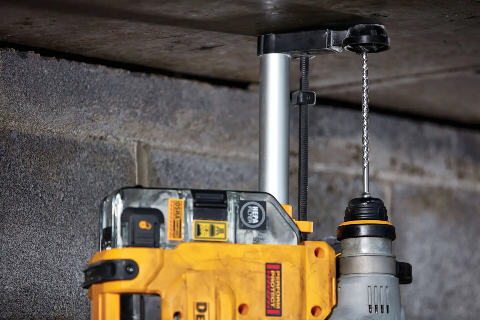A close-up of a DEWALT power drill with a drill bit attached, being used to drill into a concrete surface overhead. The tool is shown with a dust extraction attachment, highlighting construction work on a masonry ceiling.
