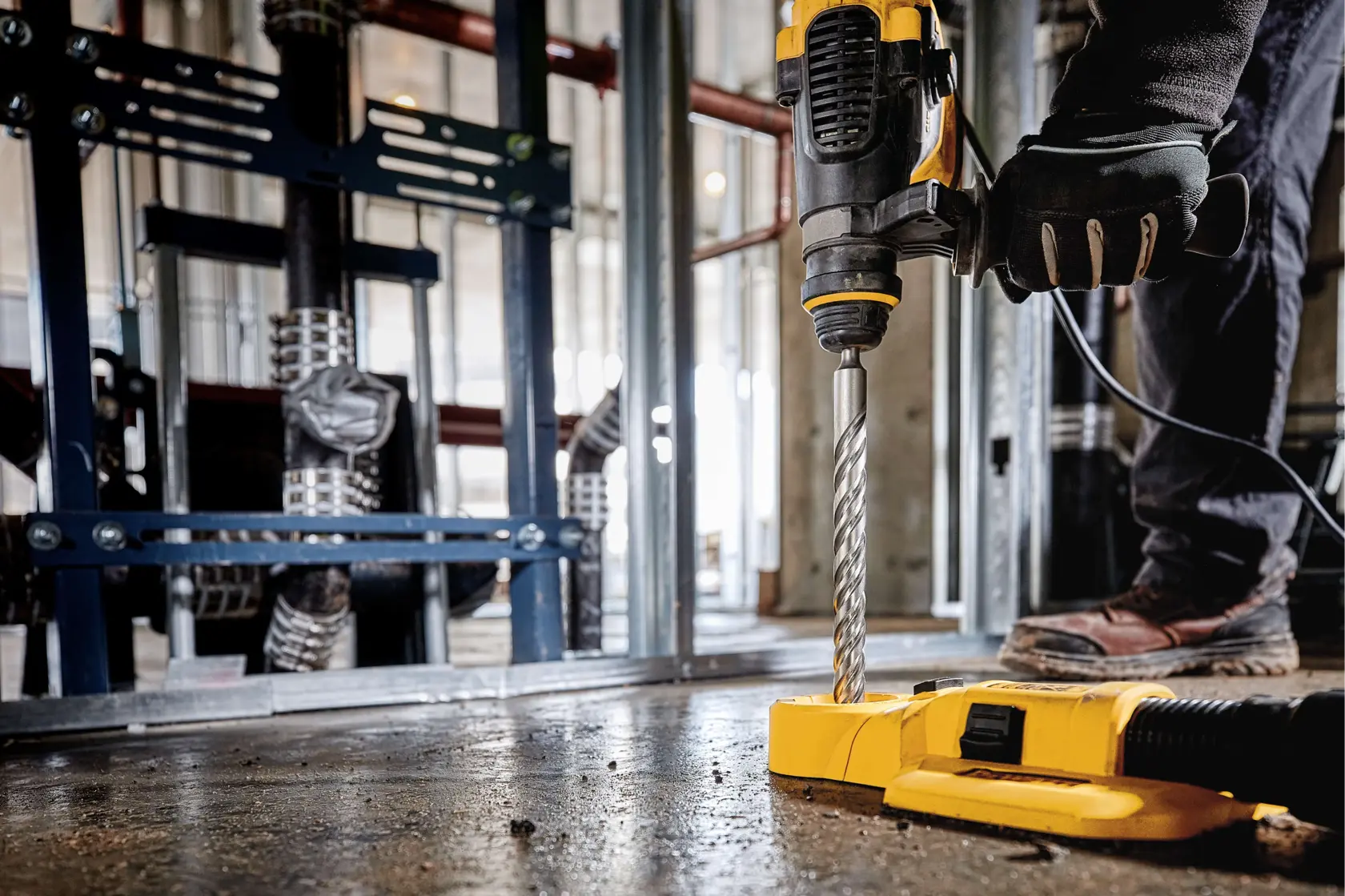 A close-up view of a worker using a DEWALT drill with a spiral drill bit to drill into the floor of an industrial or construction site. The worker is wearing gloves and boots, and the area is equipped with steel beams and pipes.