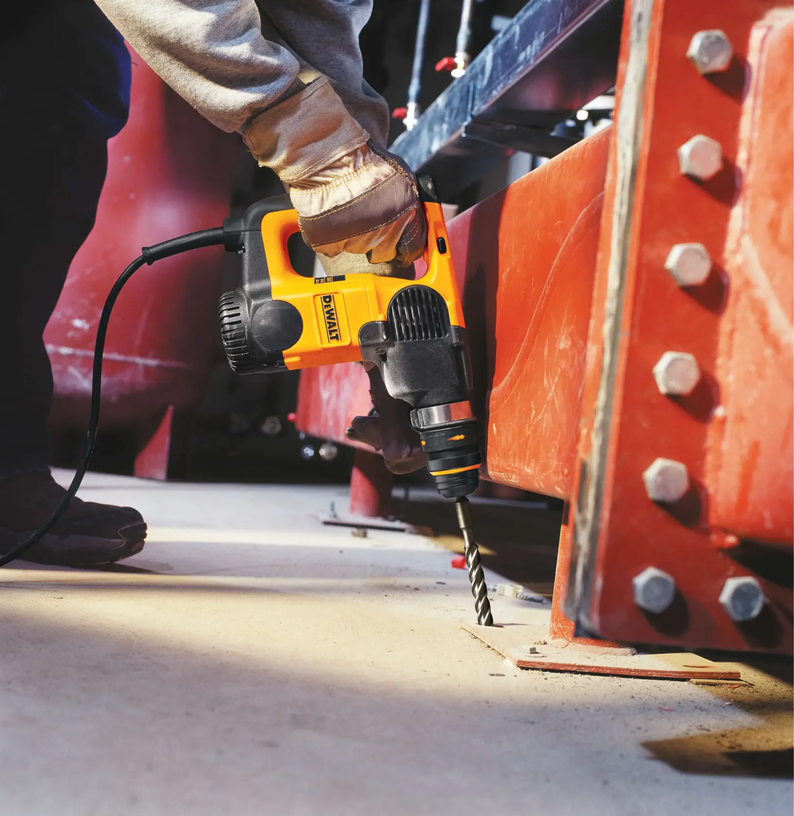 A person using a yellow and black DEWALT electric drill to drill into a metal structure, wearing work gloves and standing on a concrete floor.