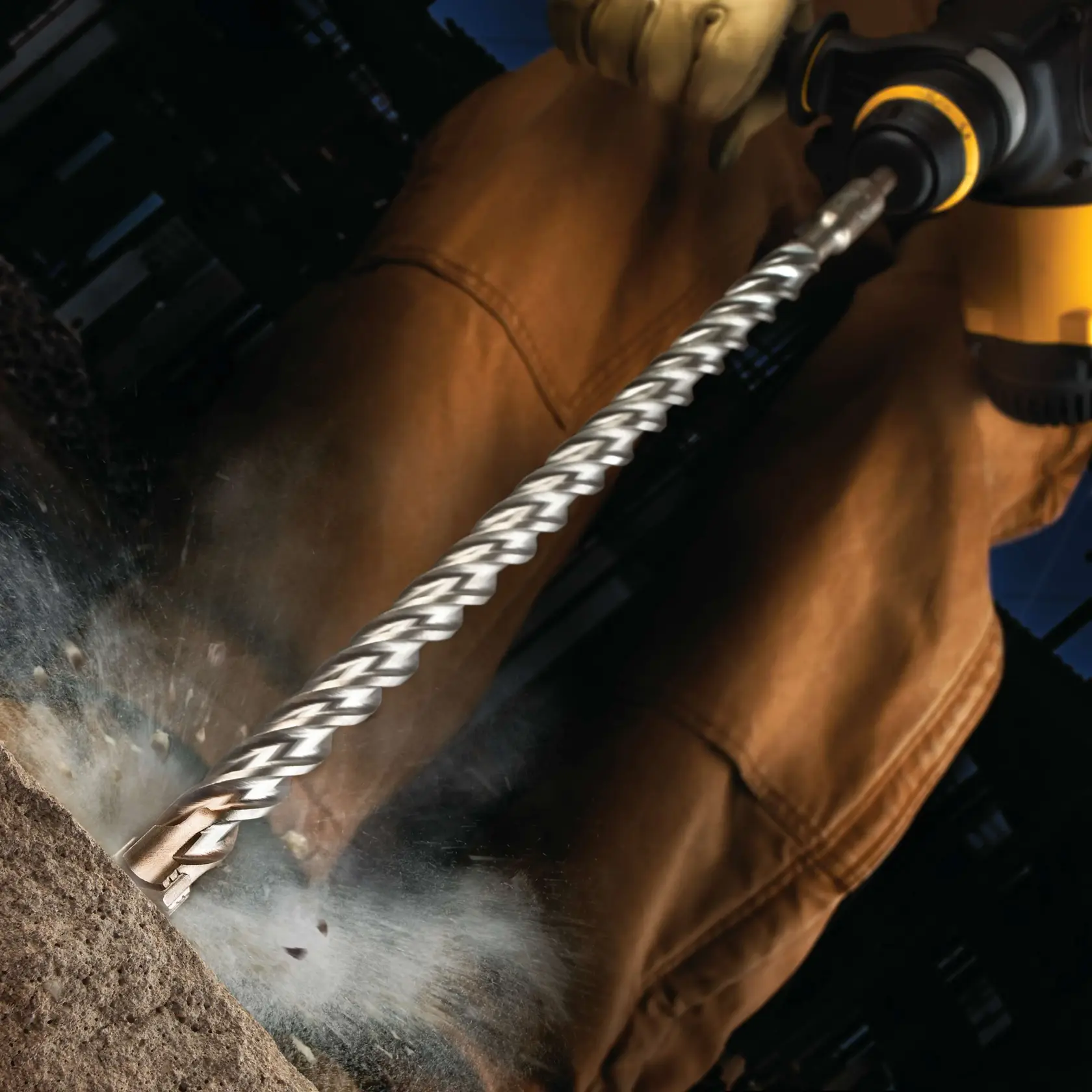 Close-up of a person using a DEWALT rotary hammer drill with a spiral bit, drilling into concrete with dust and debris flying.