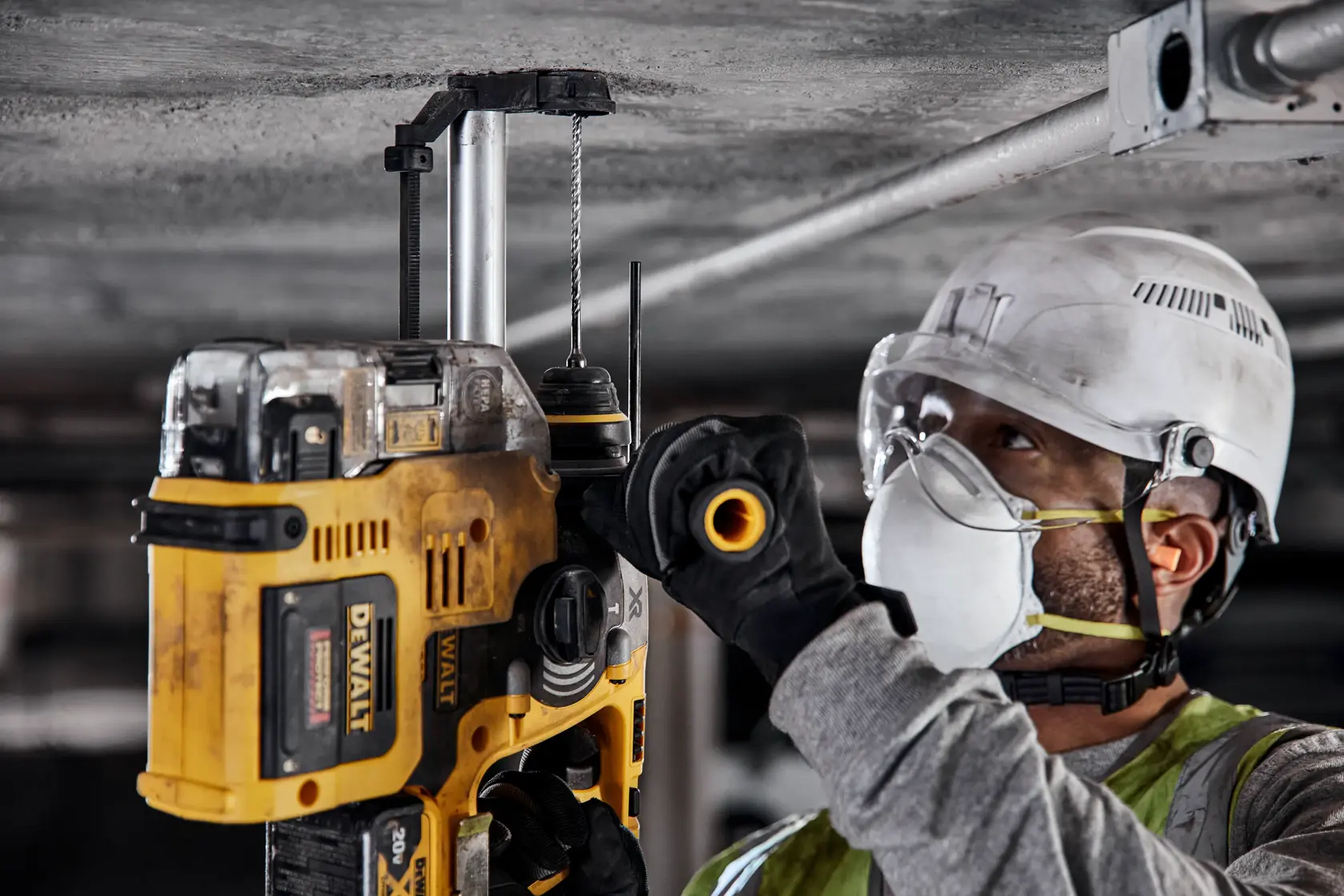 A worker wearing safety gear, including a helmet, mask, goggles, and gloves, operates a yellow DEWALT power tool to drill into a concrete ceiling.