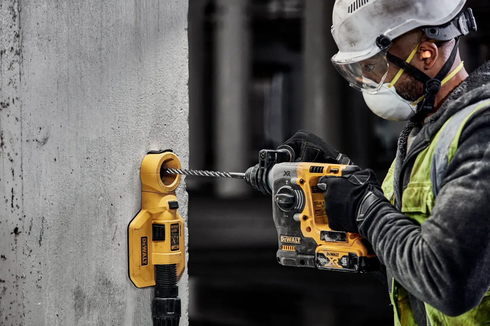 A construction worker wearing protective gear is using a DEWALT rotary hammer drill and dust extractor to drill into a concrete wall.