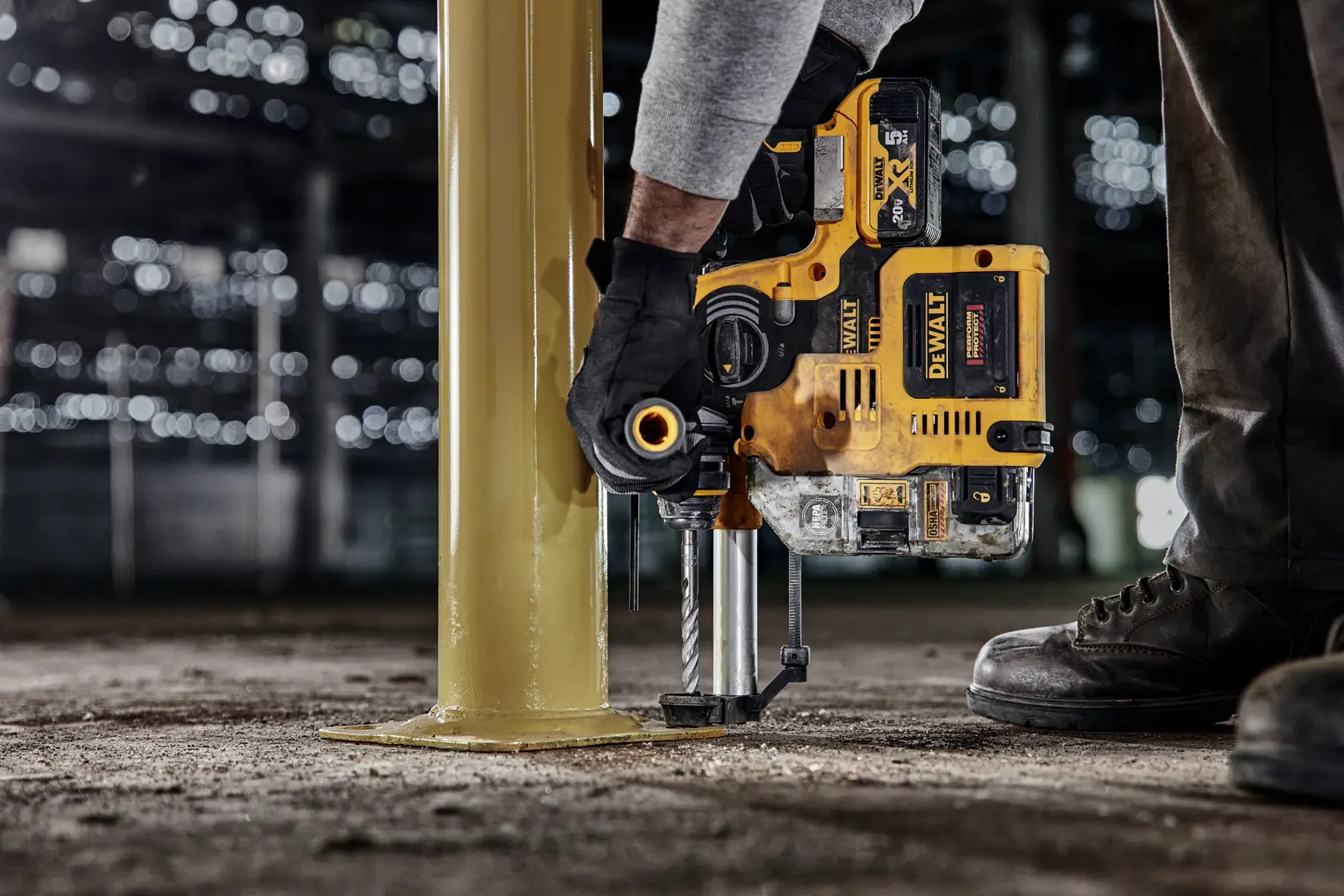 A person using a DEWALT power drill to drill into the base of a large yellow metal column, wearing gloves and work boots, in an industrial setting.