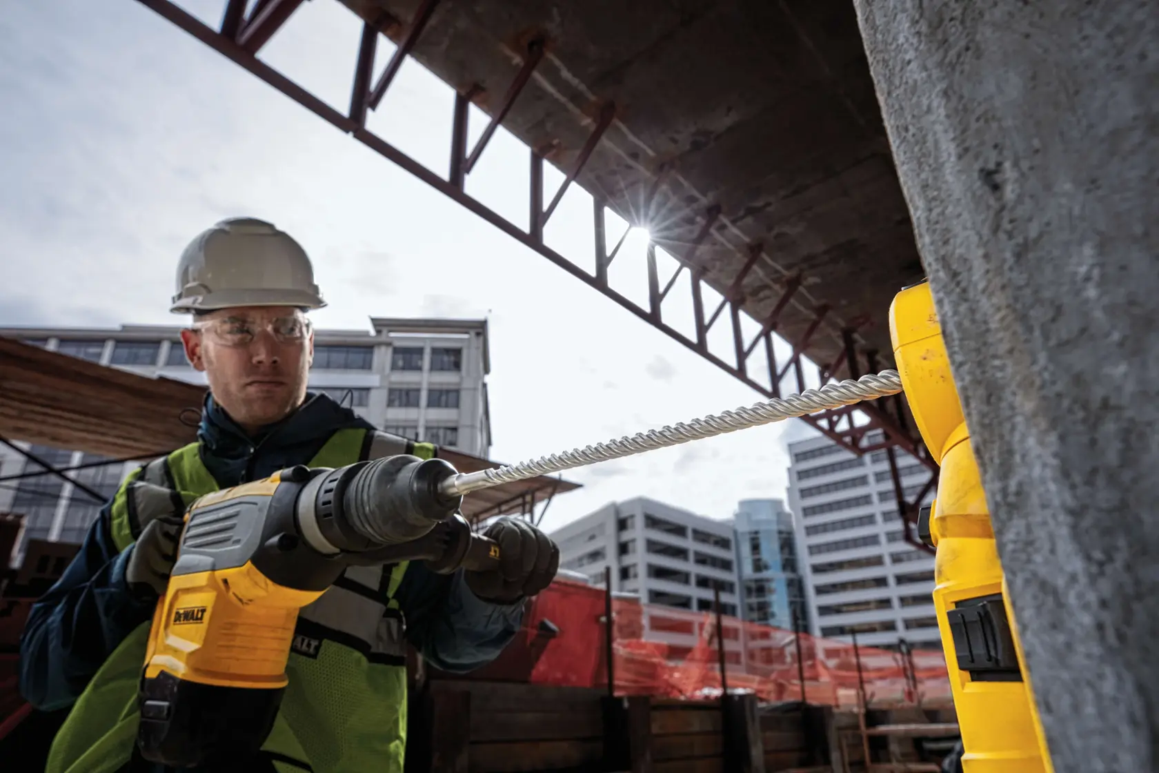 A construction worker wearing a hard hat and safety vest is using a DEWALT power tool with a long drill bit to drill into concrete at a construction site, surrounded by buildings and an unfinished structure.