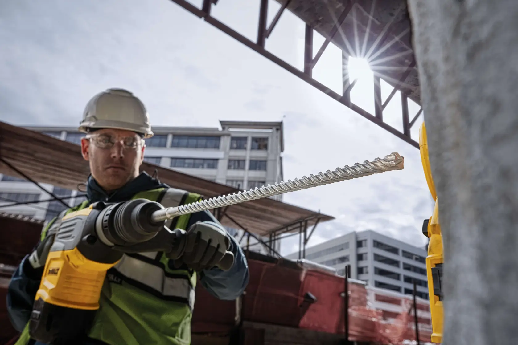 A construction worker wearing a helmet and safety vest is holding a DEWALT rotary hammer drill with a long drill bit, working at an outdoor construction site with modern buildings in the background.