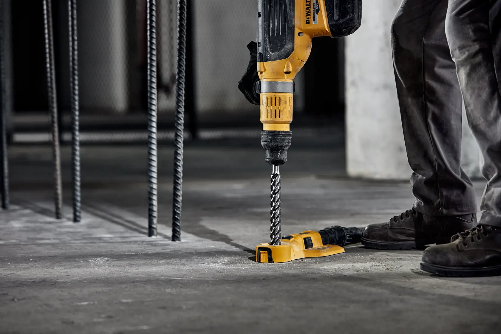 A person uses a yellow DEWALT rotary hammer drill with a spiral drill bit to drill into a concrete floor, with reinforcing steel bars nearby in an industrial setting.