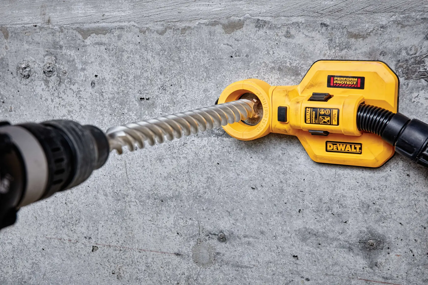 A DEWALT branded yellow dust extractor attachment is being used with a power drill on a concrete wall. The extractor is connected to a hose and surrounds the drill bit to capture dust while drilling.