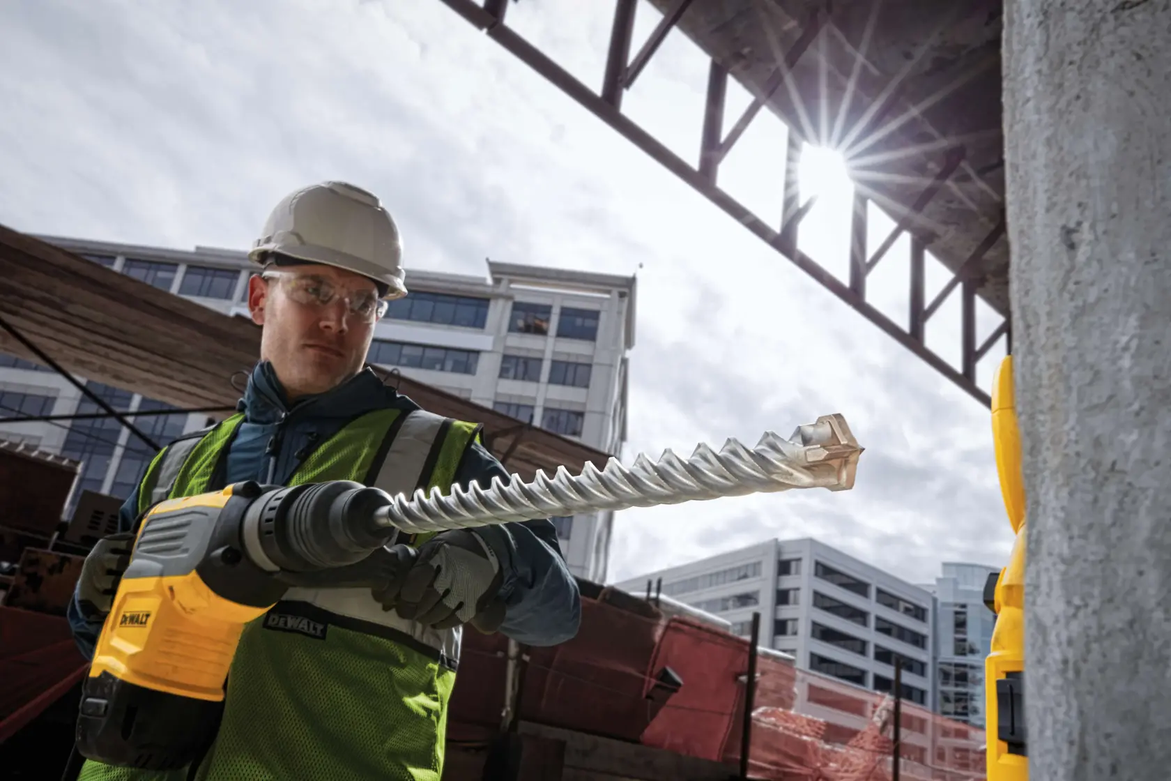 A construction worker wearing a safety helmet and vest is operating a DEWALT rotary hammer drill with a large spiral drill bit at an urban job site. Buildings and construction materials are visible in the background.
