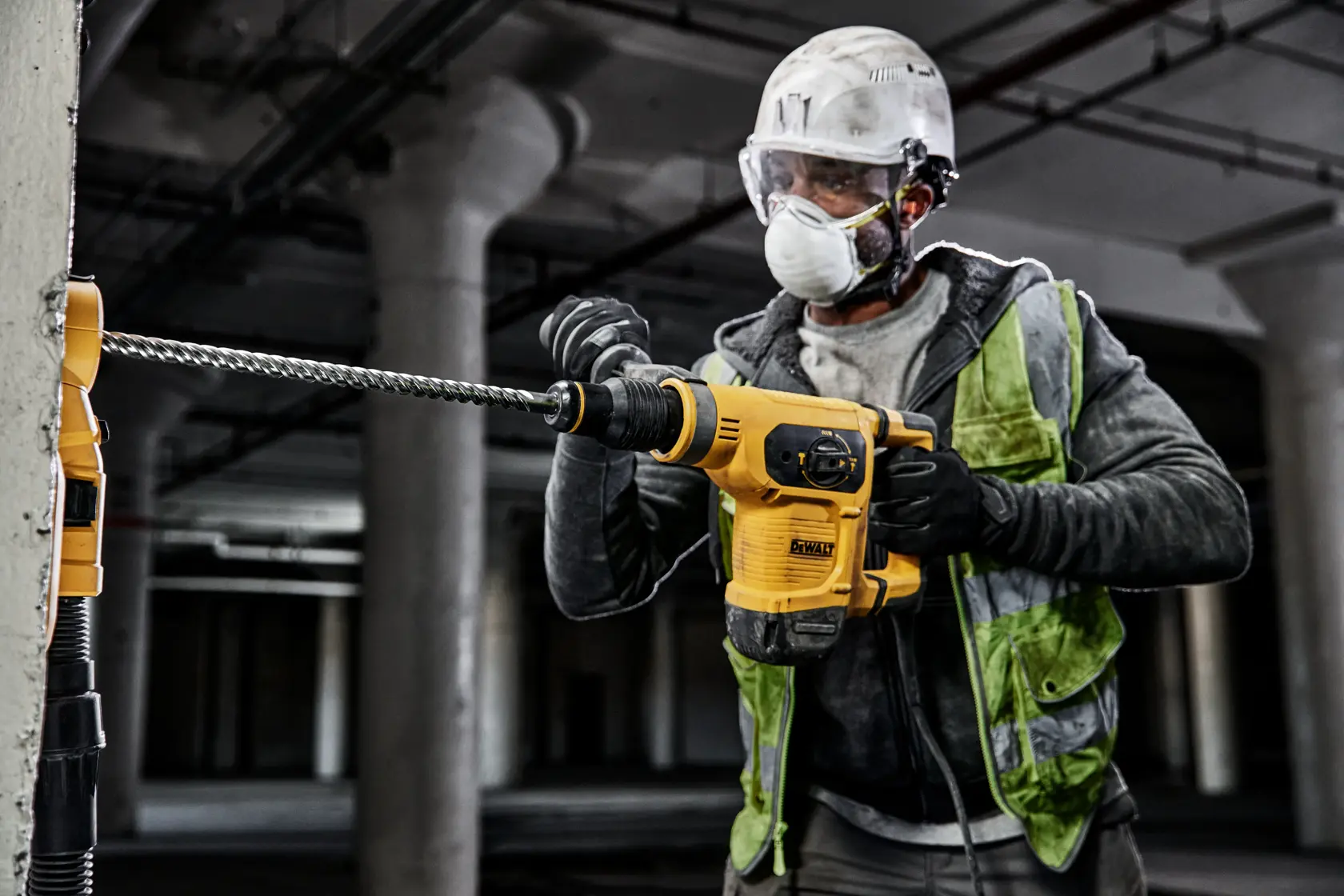 A construction worker operating a yellow DEWALT DW5824 rotary hammer drill, drilling into a concrete wall in an industrial setting. The worker is wearing a safety helmet, high-visibility vest, gloves, and protective eyewear.
