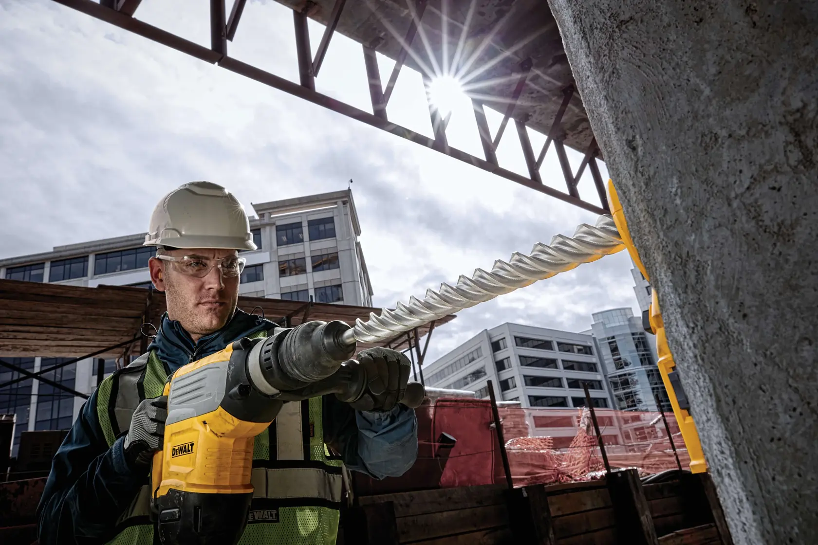A construction worker using a DEWALT rotary hammer drill with a long, spiral bit to drill into a concrete pillar at a construction site. The worker is wearing safety gear, including a helmet and reflective vest, and modern buildings are visible in the background.