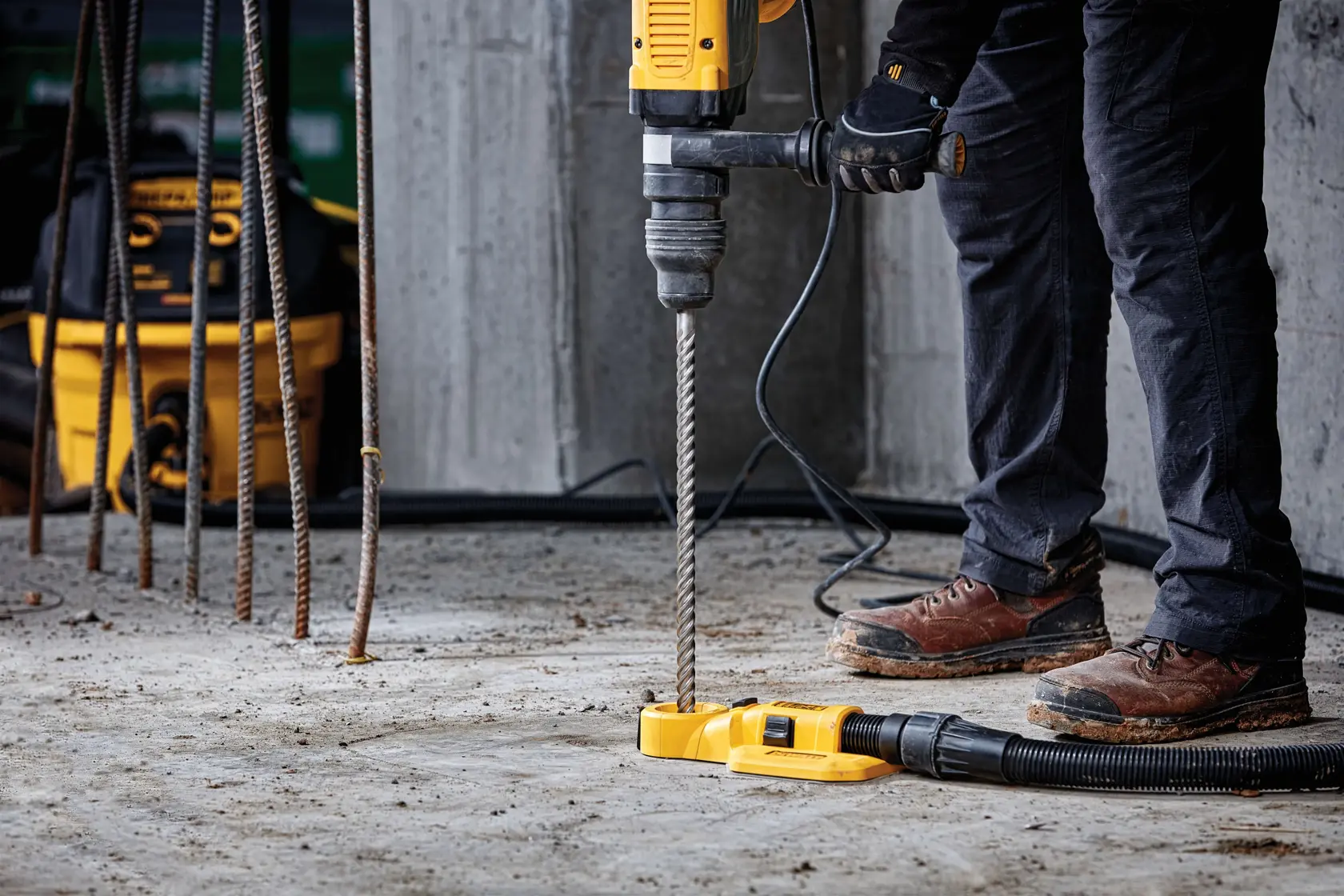 A person using a DEWALT rotary hammer drill with a long drill bit to drill into a concrete floor. The person is wearing black pants, brown work boots, and gloves. There is a dust collection attachment connected to a vacuum hose. Several steel rods and a DEWALT tool bag are visible in the background.