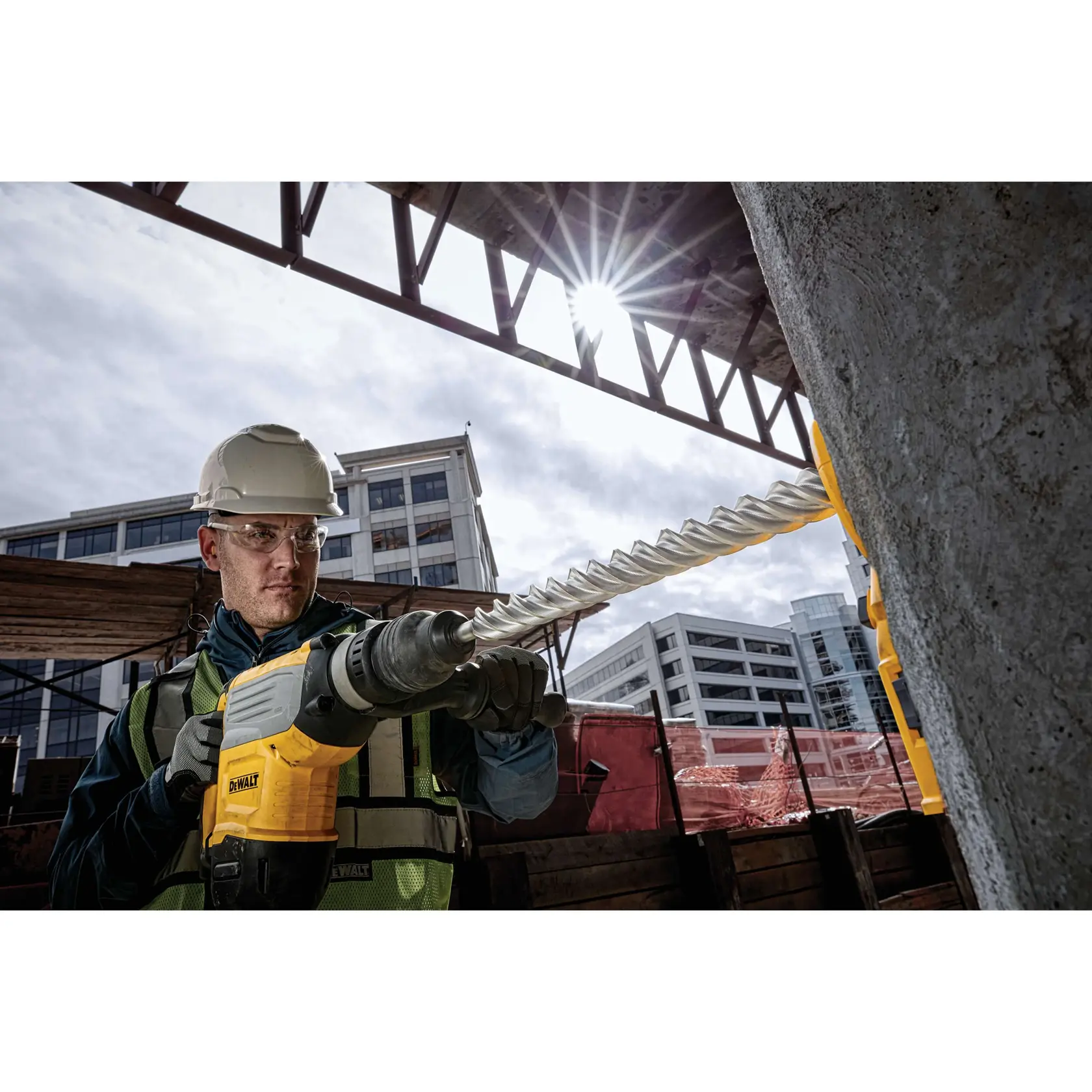 A construction worker using a DEWALT power tool with a long drill bit to drill into concrete at an outdoor construction site. The worker is wearing safety gear including a helmet and reflective vest. Modern buildings are visible in the background.