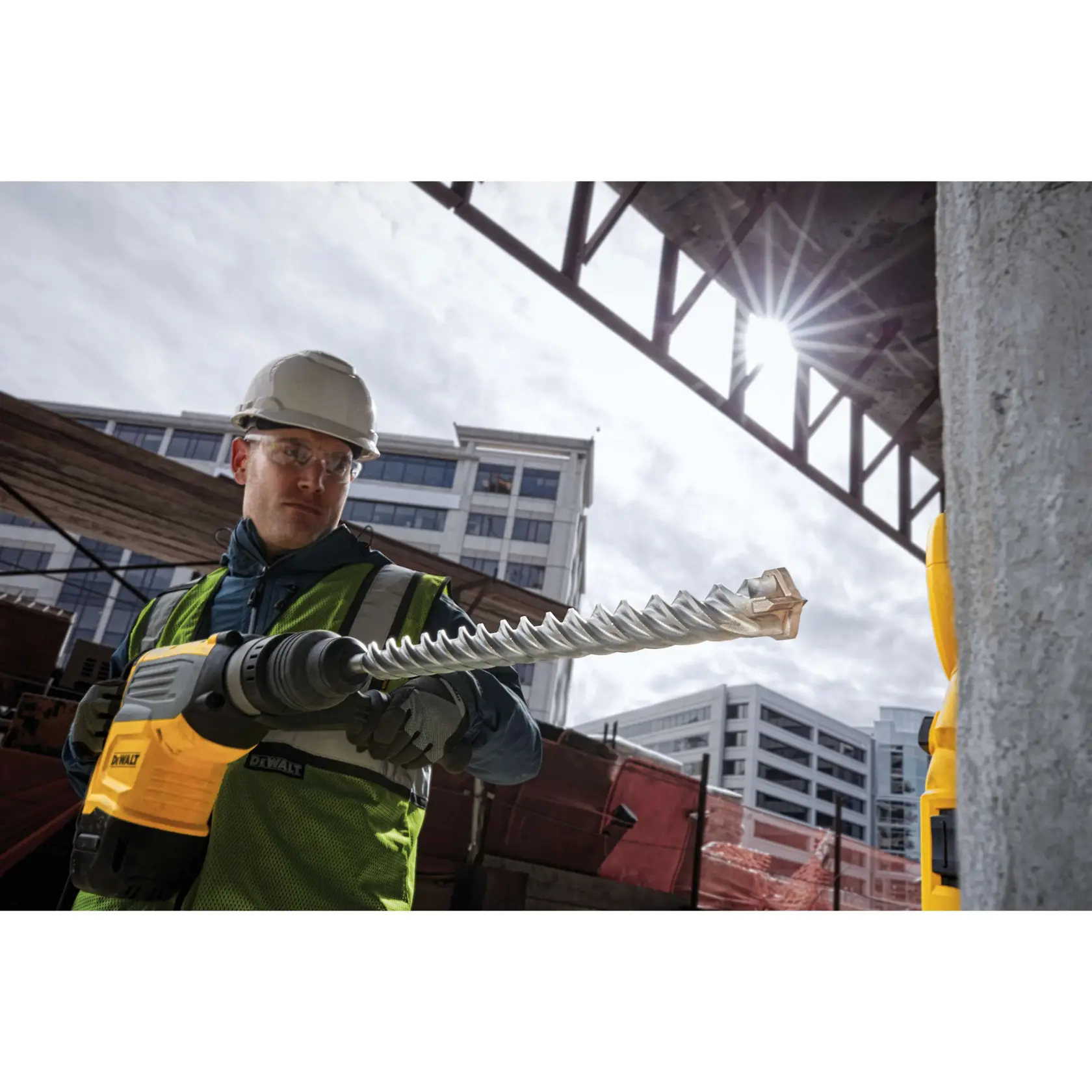A worker wearing a safety vest and hard hat is using a DEWALT power tool with a large drill bit at a construction site in front of modern office buildings.
