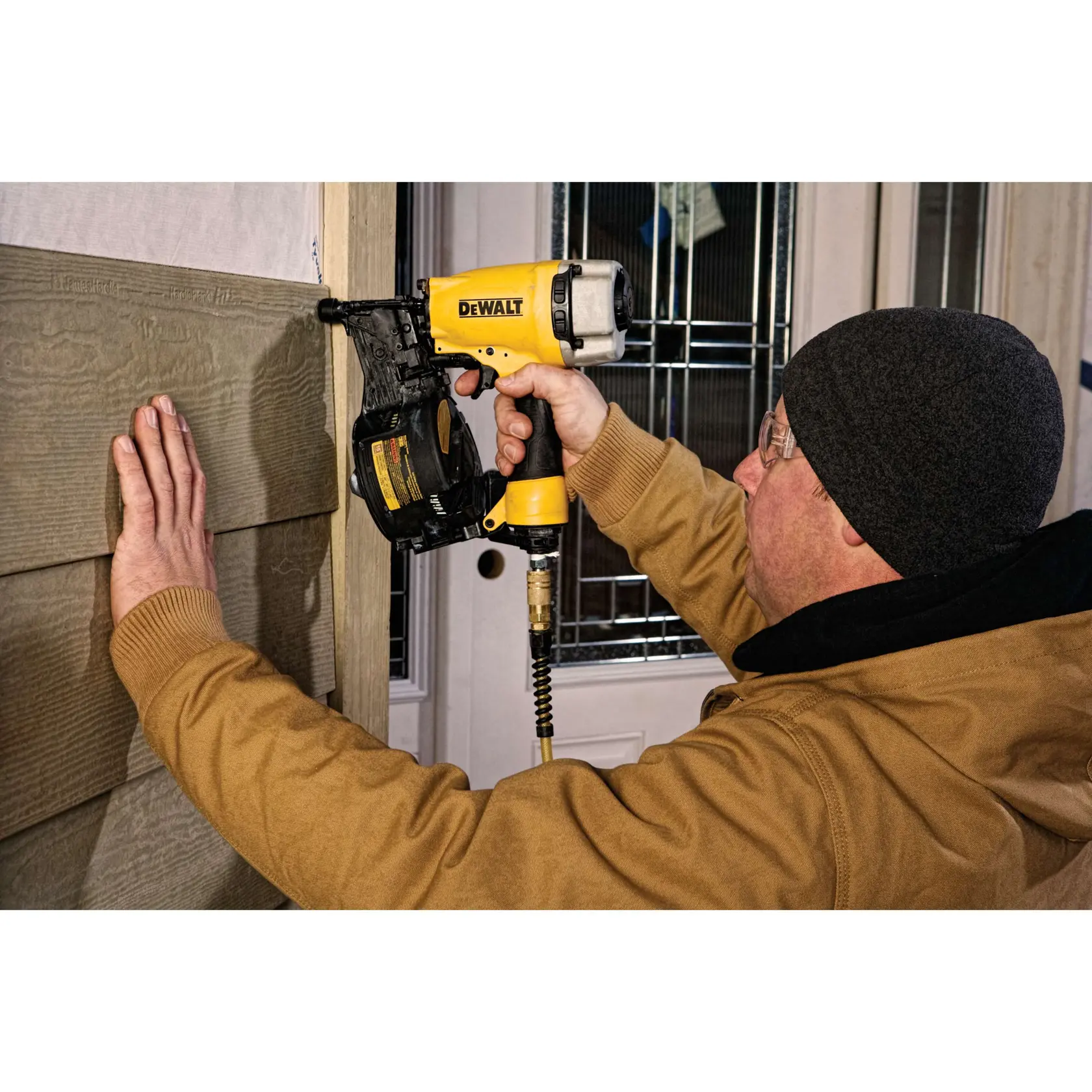 A person in a brown jacket uses a DEWALT DW66C-1 pneumatic coil siding nailer to install siding on a wall. The person’s face is blurred for privacy.