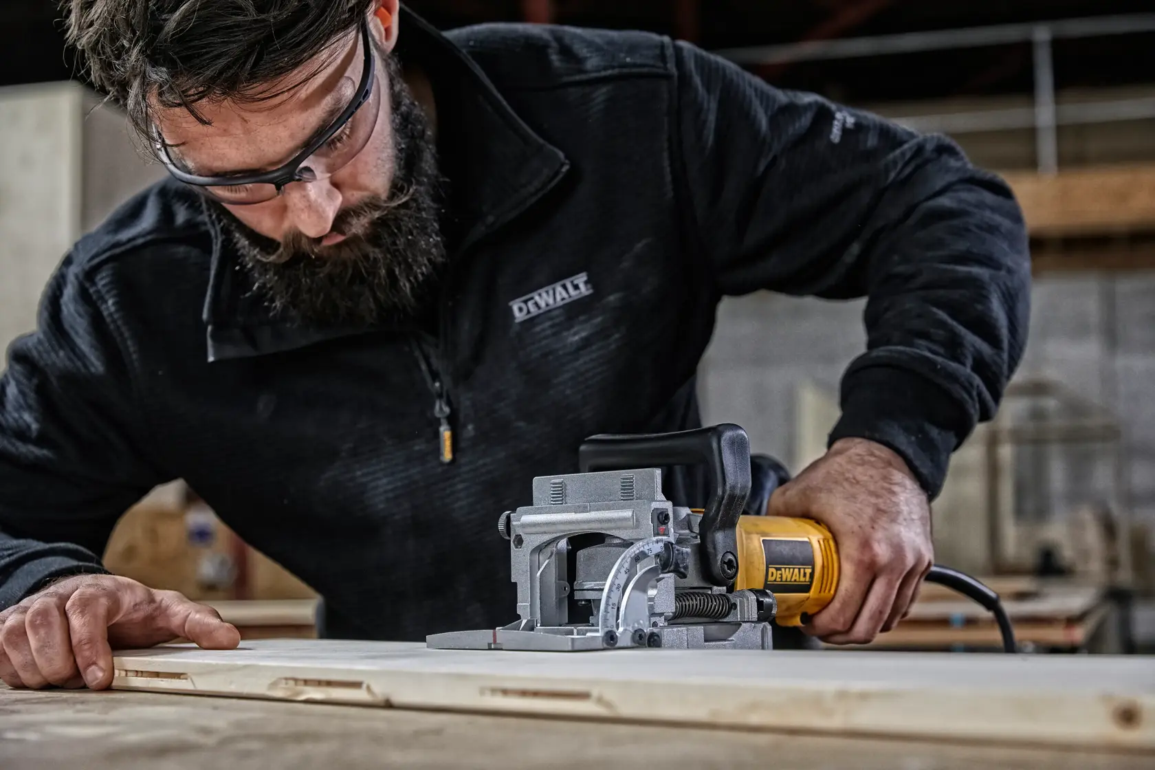 A person using a DEWALT DW682K biscuit joiner on a wooden surface in a workshop. The user's face is blurred, and they are wearing a black DEWALT jacket.