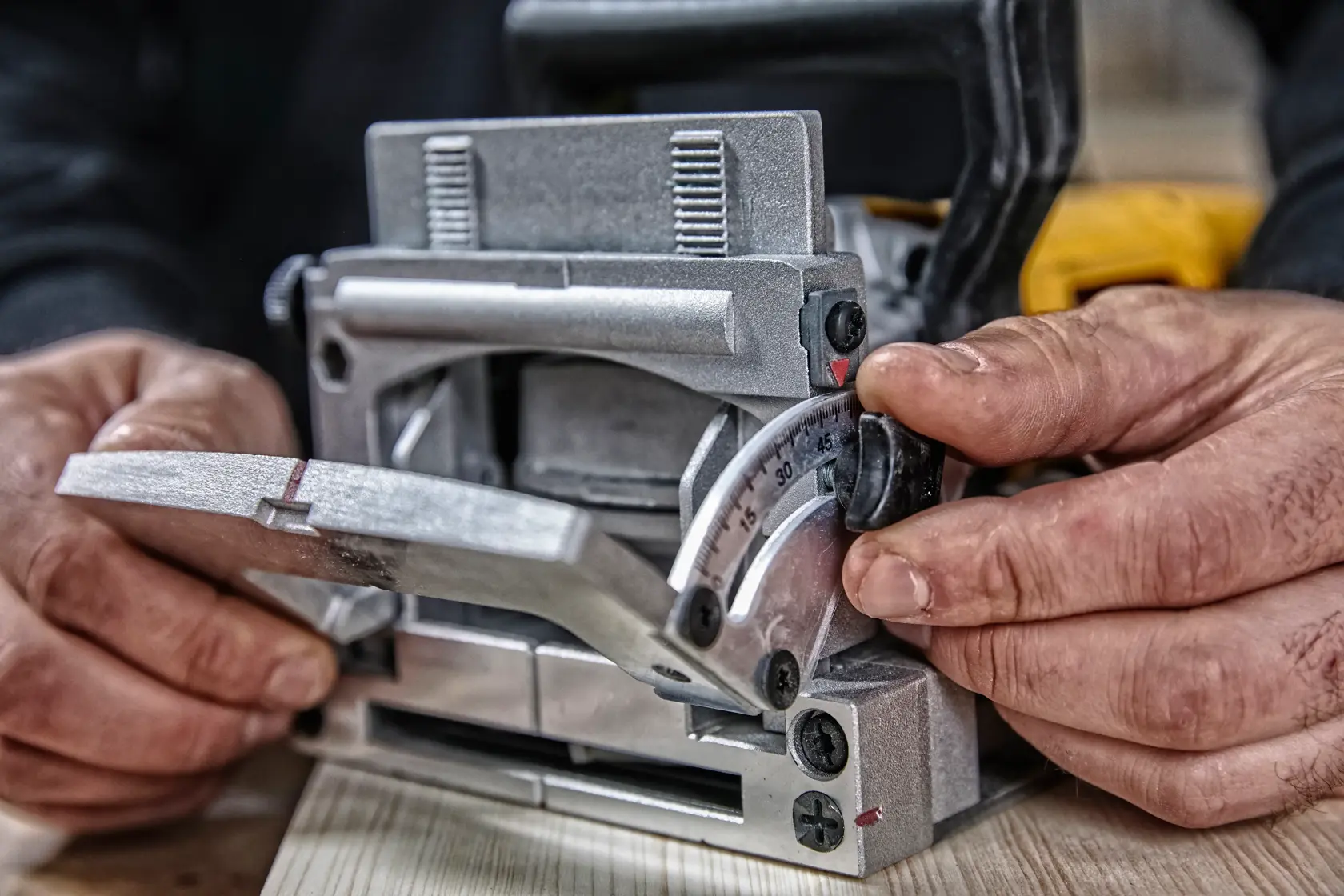 Close-up of hands adjusting the angle settings on a DEWALT biscuit joiner tool for woodworking, with the tool resting on a wooden plank.
