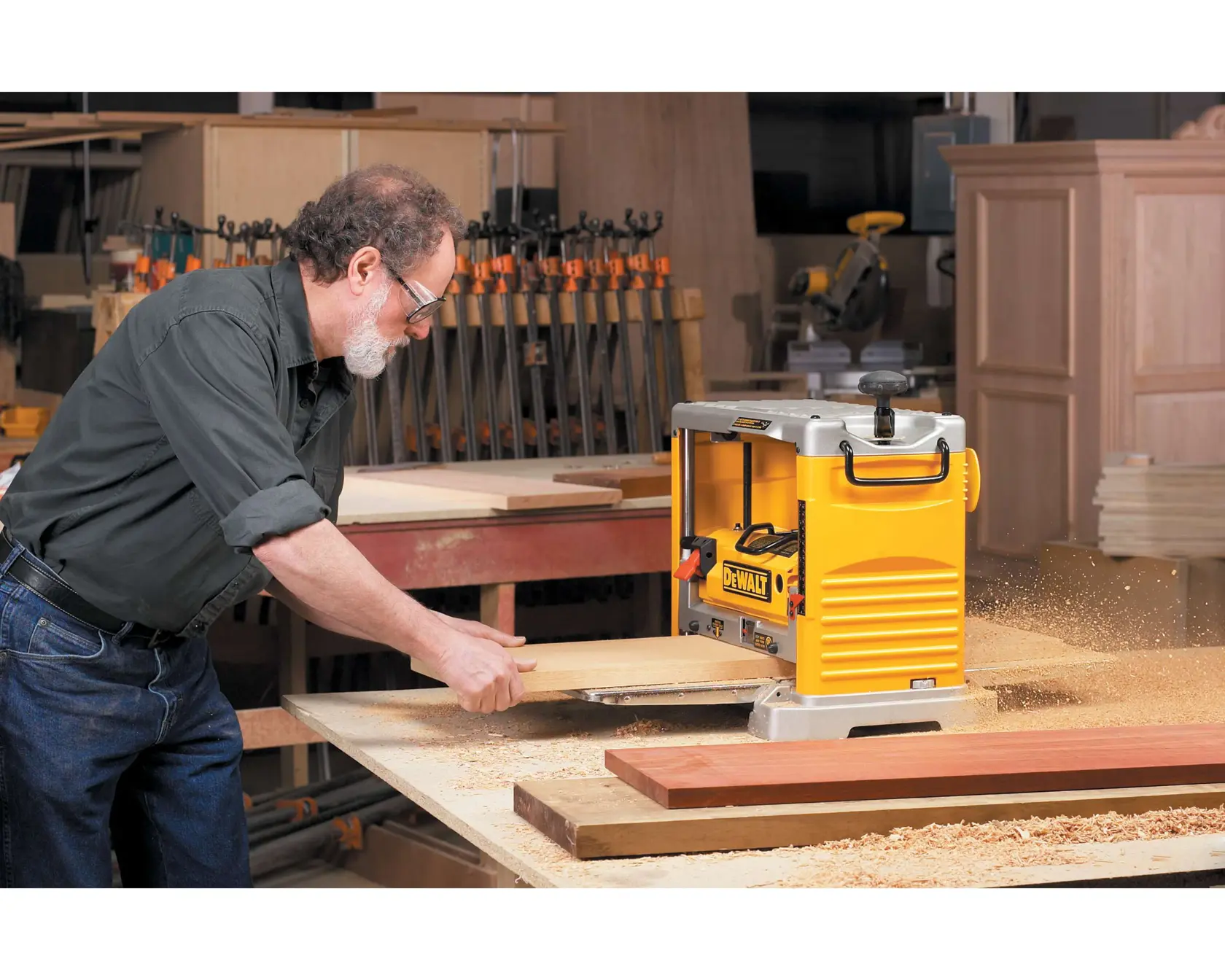 A person operating a DEWALT DW734 wood planer in a workshop, smoothing a piece of wood with wood shavings flying around. The workspace is equipped with clamps, tables, and wooden boards.