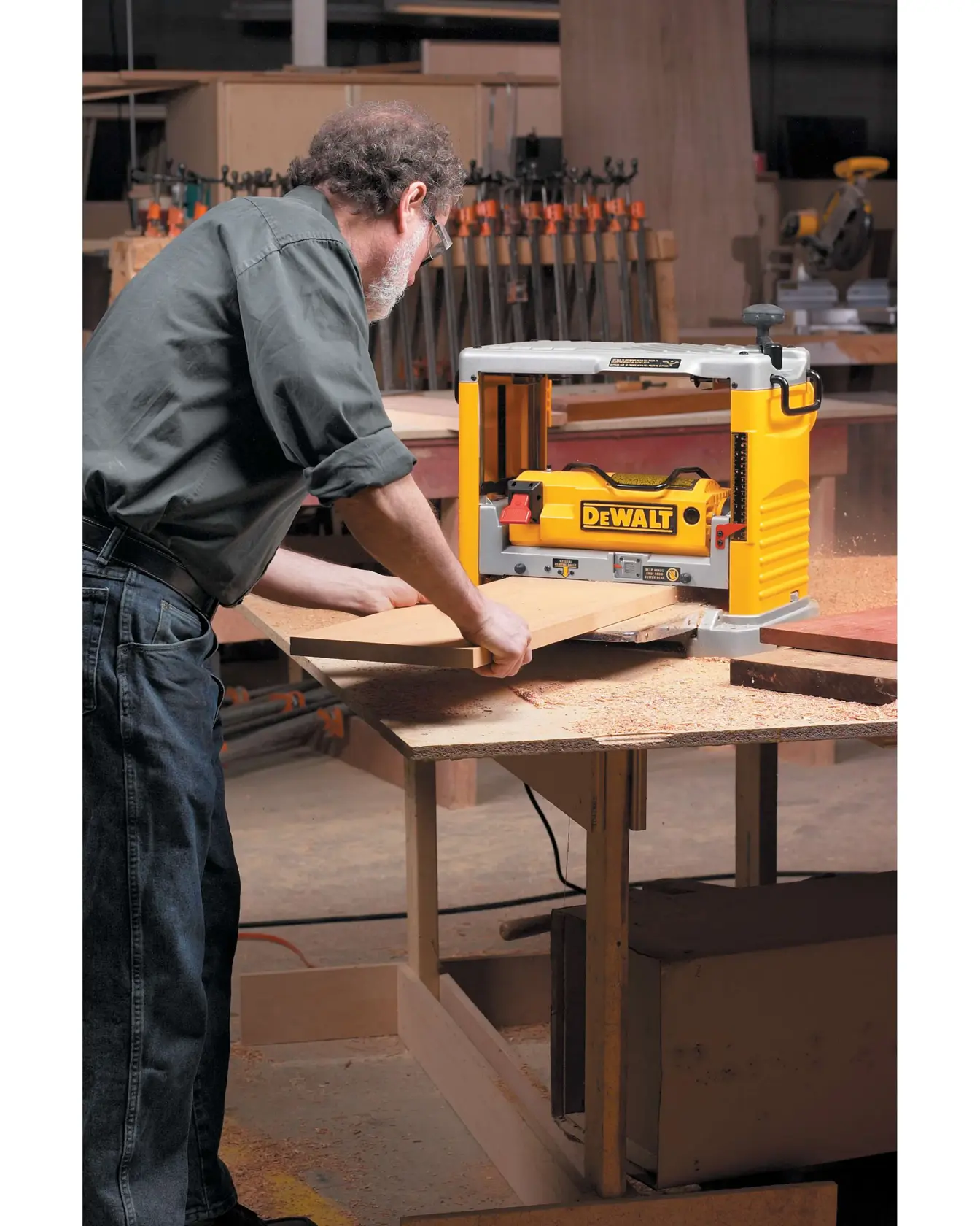 A person using a DEWALT DW734 wood planer on a workbench in a woodworking workshop. The planer is being used to smooth a wooden board, with wood shavings visible on the table.