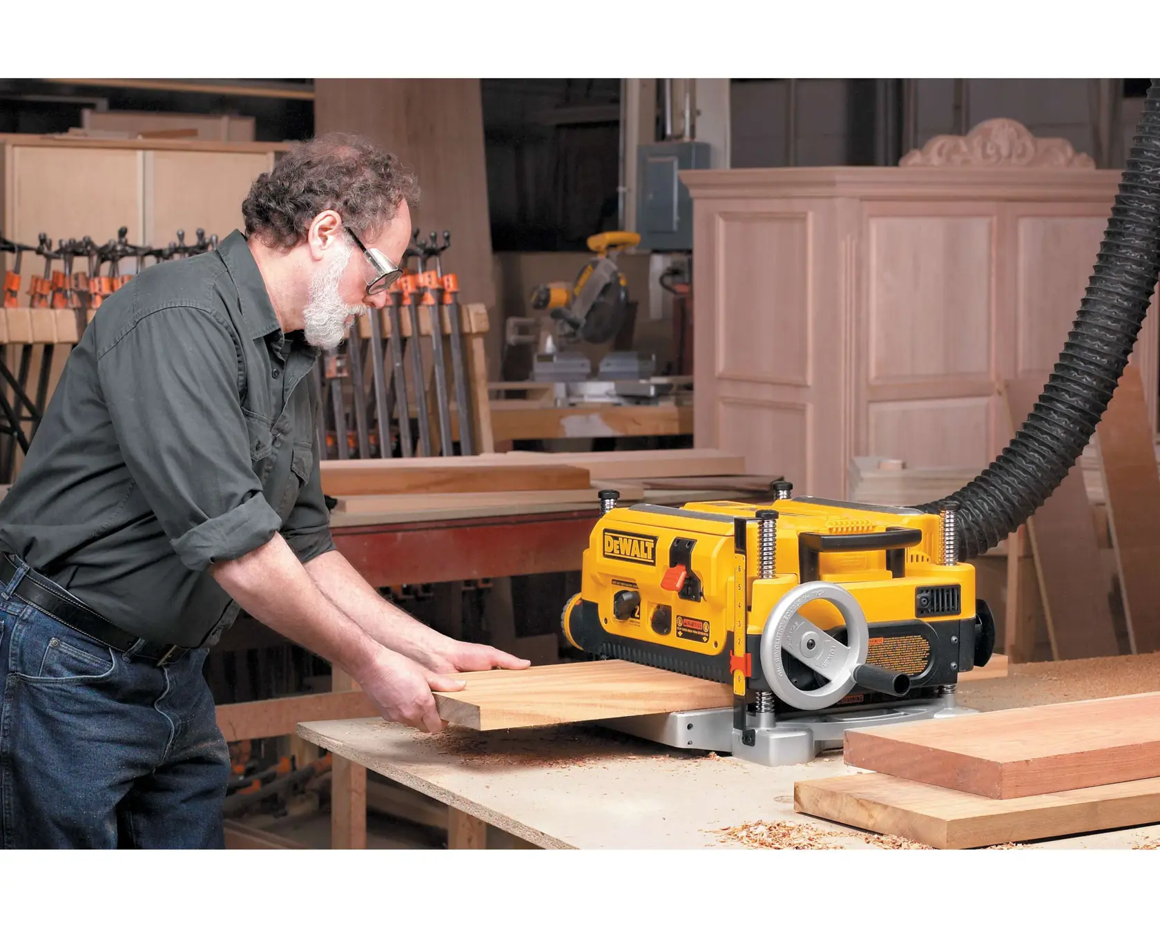 A person feeds a wooden board into a DEWALT DW735 planer machine on a workbench in a woodworking shop. The planer is yellow and black, and connected to a dust extraction hose.