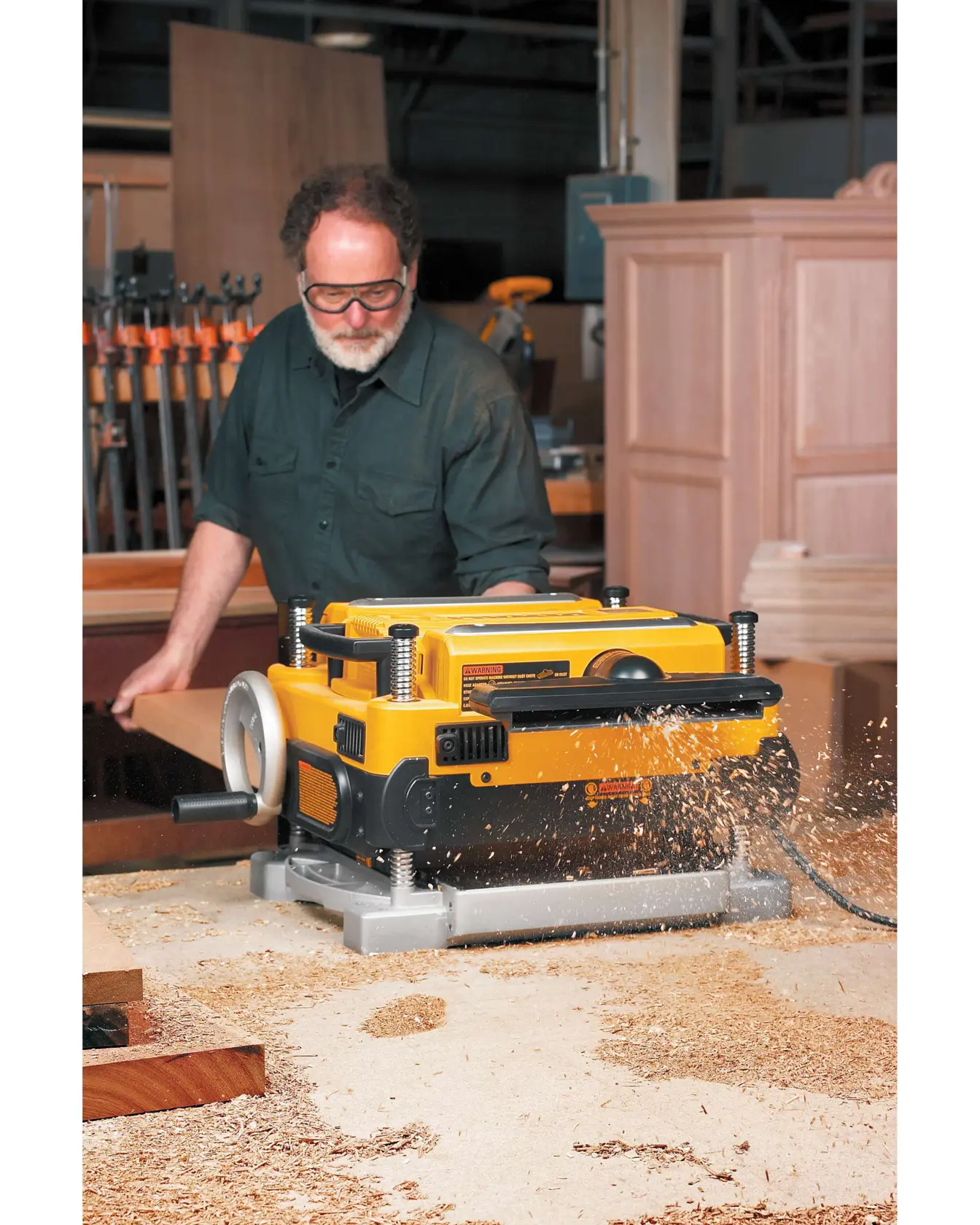 A DEWALT DW735 thickness planer in operation, with wood shavings being ejected, placed on a workbench in a woodworking workshop. A person stands behind the planer, and there are various woodworking tools and cabinets visible in the background.