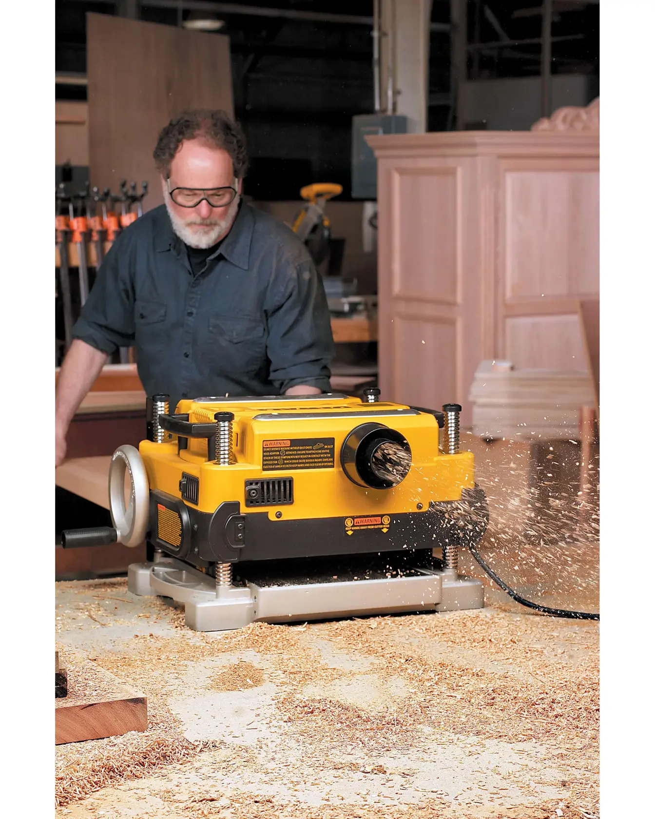 A DEWALT DW735 yellow and black wood planer being used in a workshop. Wood shavings are flying out as the machine operates, with a blurred person standing behind it. There are woodworking tools and cabinets visible in the background.