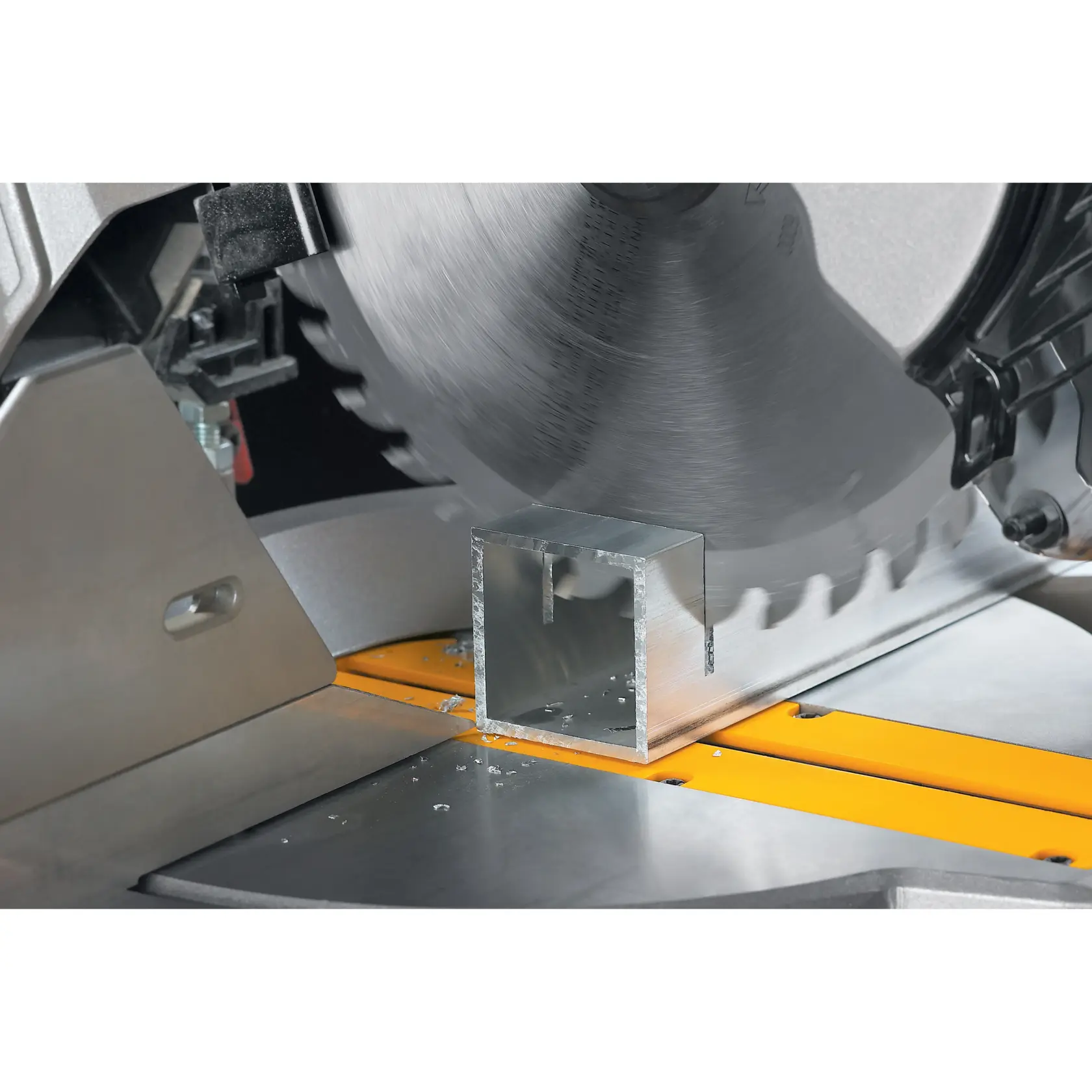 A close-up view of a circular saw cutting through a metal square tube on a workbench. Metal shavings are visible on the surface.