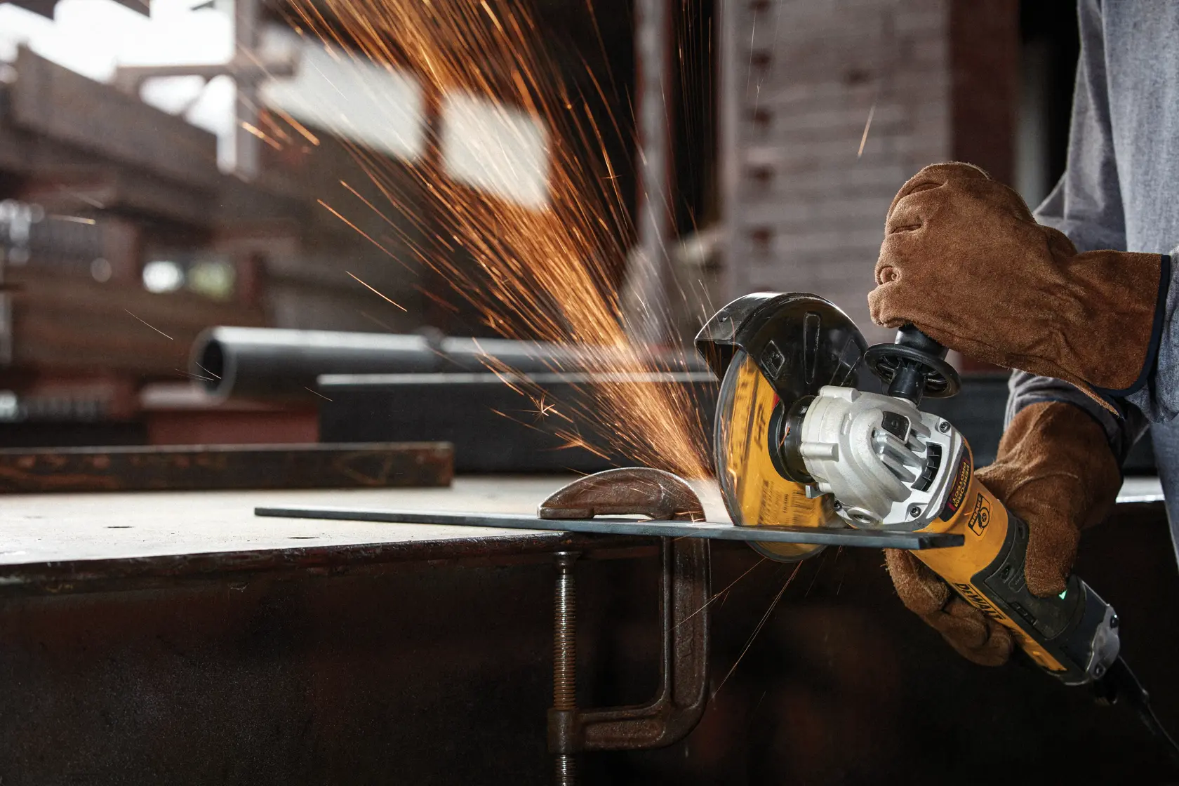 Person wearing brown protective gloves using a DEWALT angle grinder to cut metal, with sparks flying, in an industrial workshop setting.