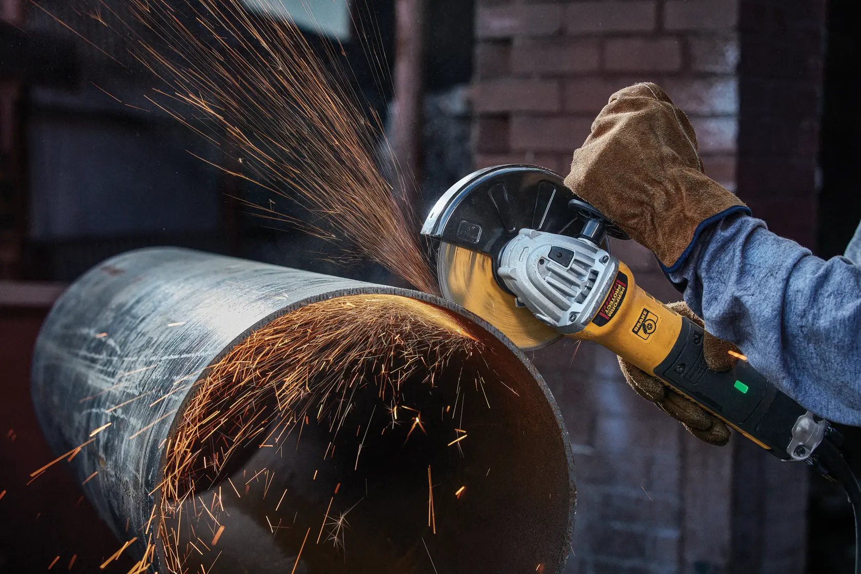 A person wearing protective gloves is using a yellow DEWALT angle grinder to cut a large metal pipe, with sparks flying from the cutting area.