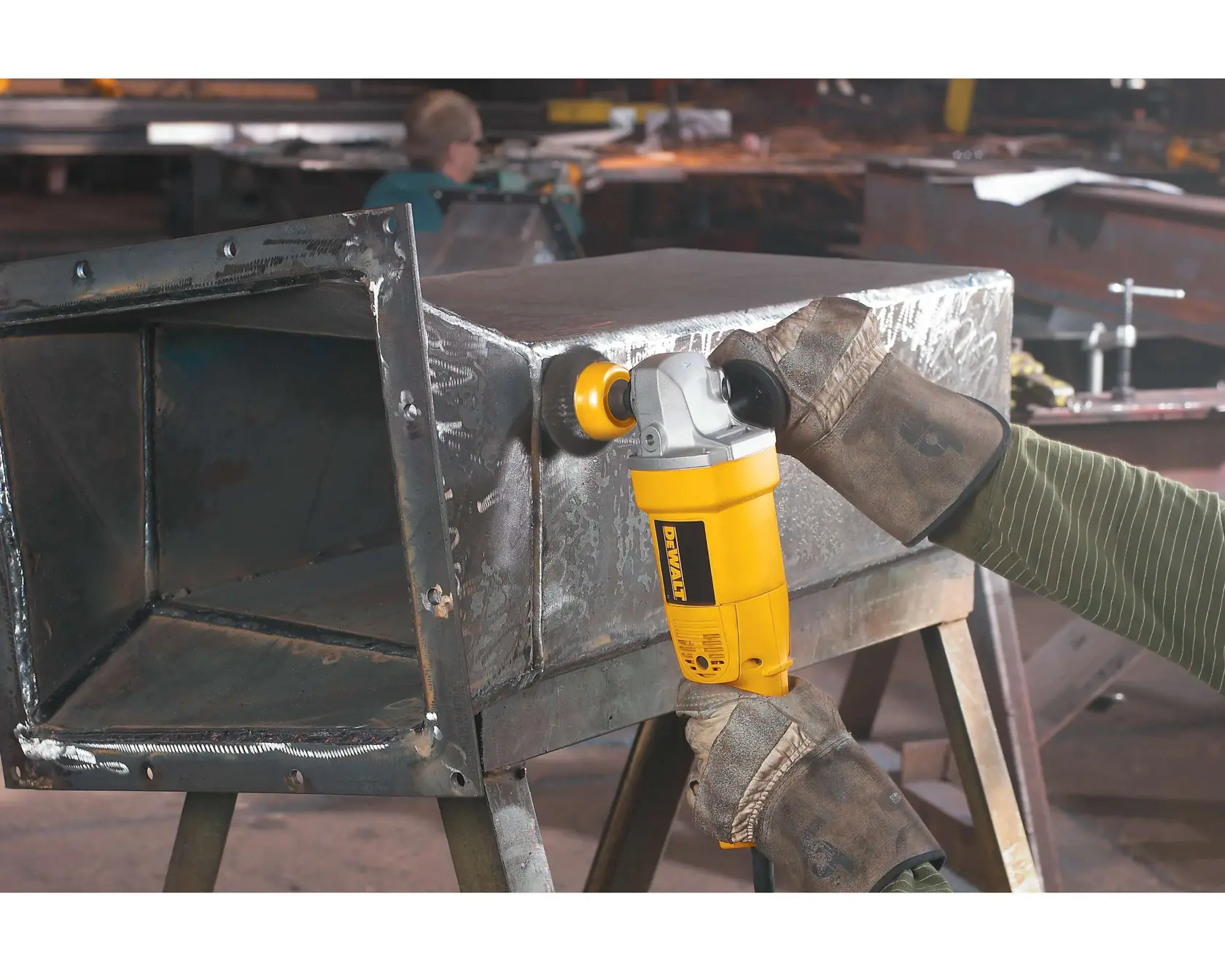 A person wearing protective gloves uses a yellow DEWALT angle grinder (model DW831) to grind the edge of a metal box in a workshop.