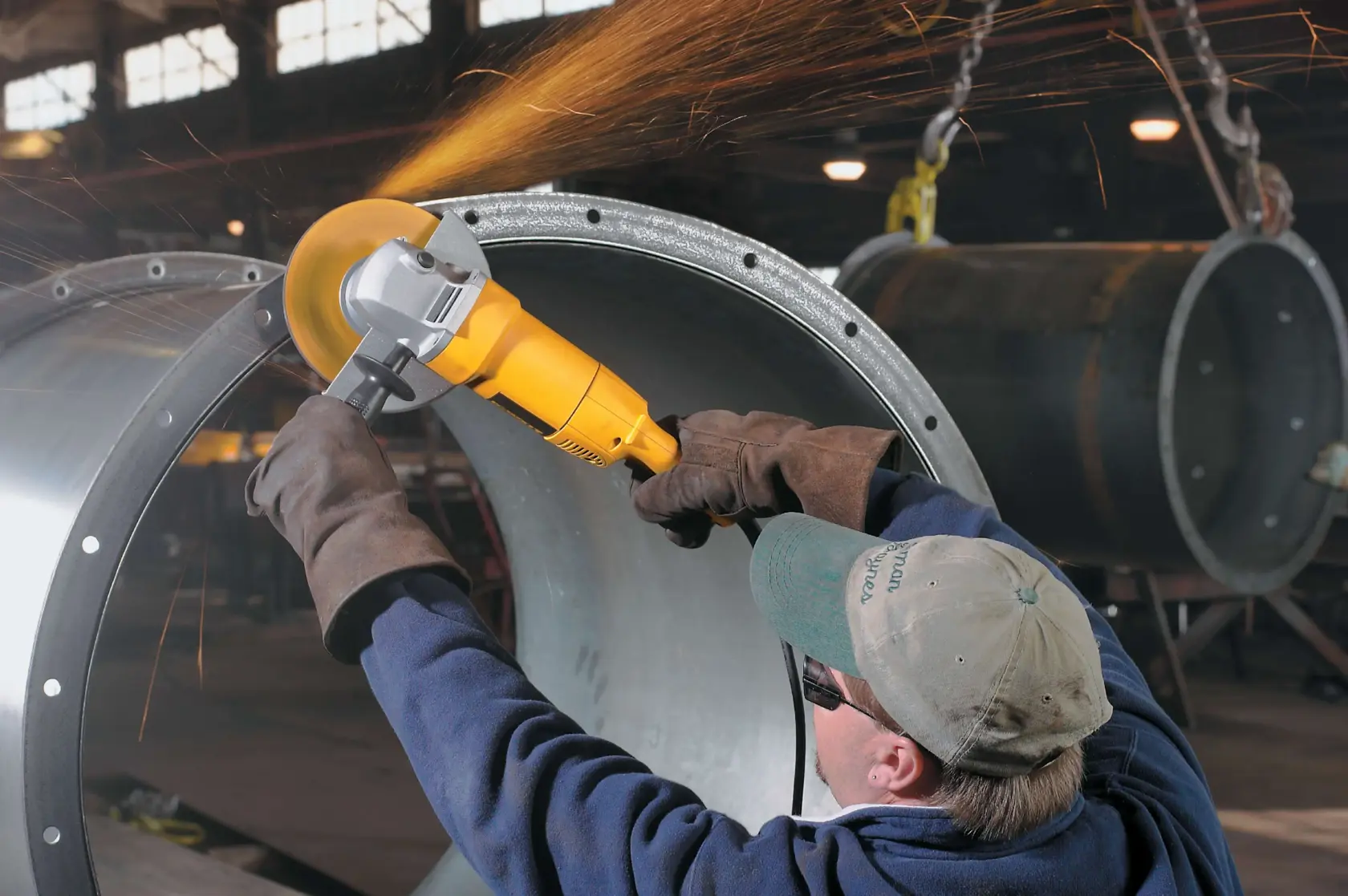 A person wearing protective gloves, a cap, and safety glasses uses a yellow DEWALT angle grinder to polish or cut the edge of a large metal cylinder, producing sparks, inside an industrial workshop.
