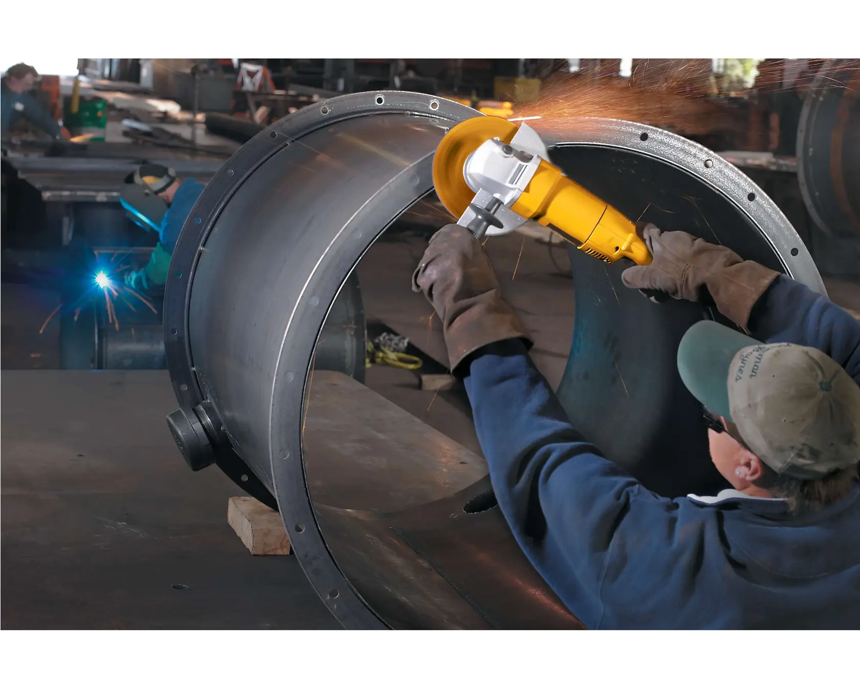Person using DEWALT grinder on large metal pipe, sparks flying, industrial setting, safety gloves and cap worn.