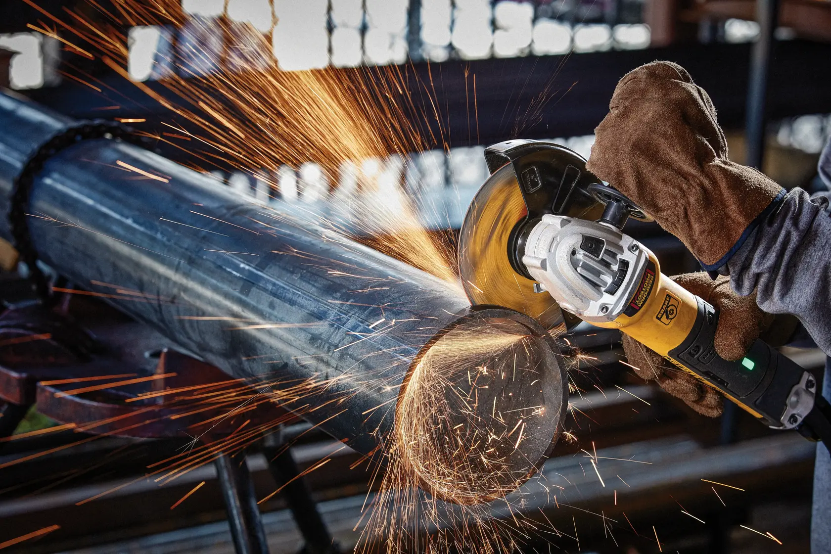 A person wearing protective gloves uses a DEWALT angle grinder to cut a large metal pipe, producing bright sparks.