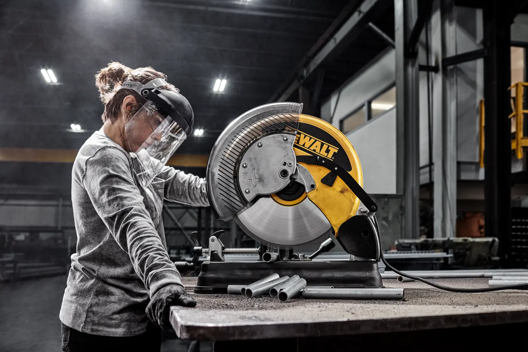 A person wearing safety gear operates a DEWALT DW872 metal cutting saw in an industrial workshop, cutting metal pipes on a worktable.