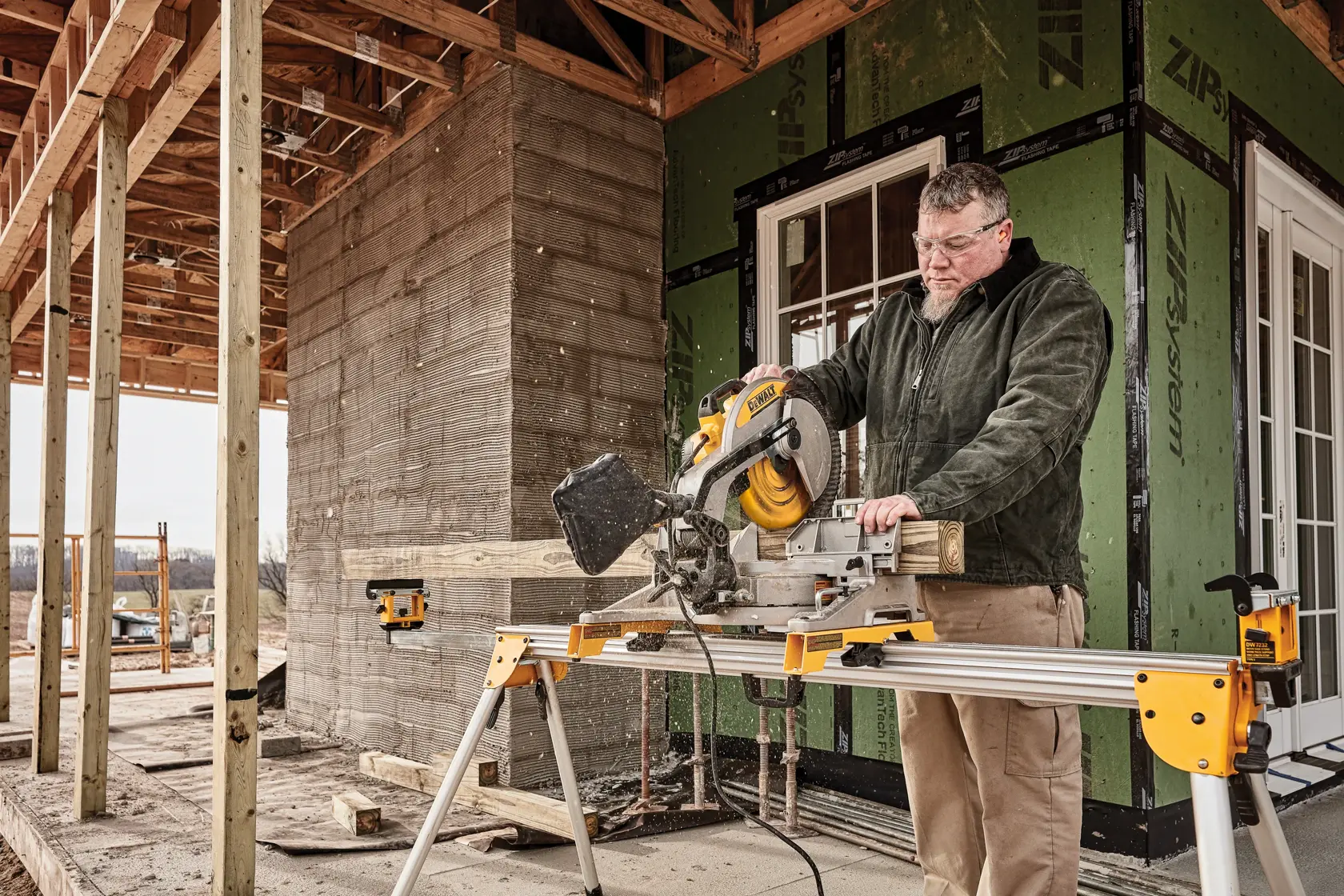 A person using a DEWALT miter saw to cut wood on a construction site. The saw is mounted on a portable stand, and the surroundings show a partially constructed building.