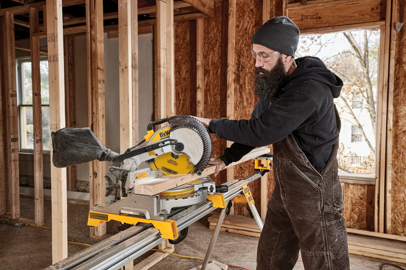 A person wearing work overalls and a beanie is using a DEWALT miter saw to cut a piece of wood inside a building under construction. The surroundings feature exposed wooden framing and unfinished walls.