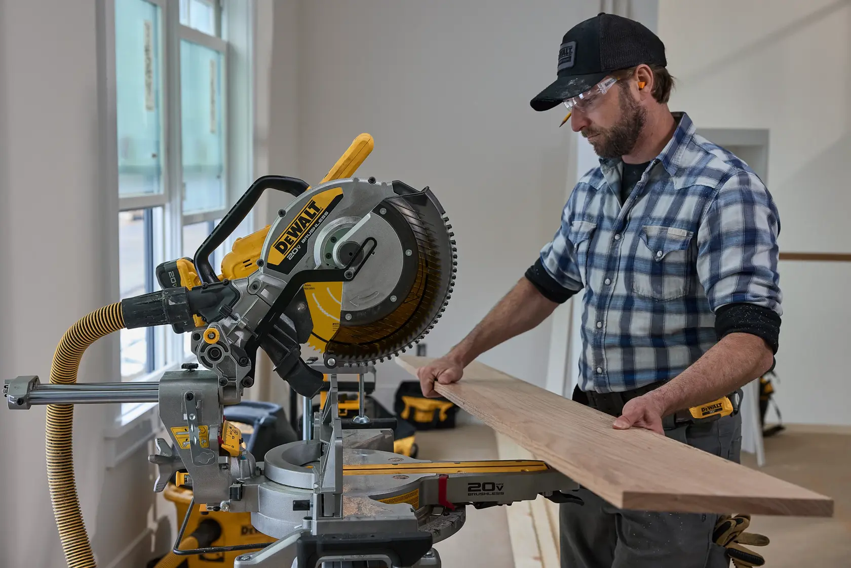 A person using a DEWALT miter saw to cut a piece of wood inside a workshop. The person’s face is blurred and they are wearing a plaid shirt and a cap. The saw is yellow and silver, with a vacuum hose attached.