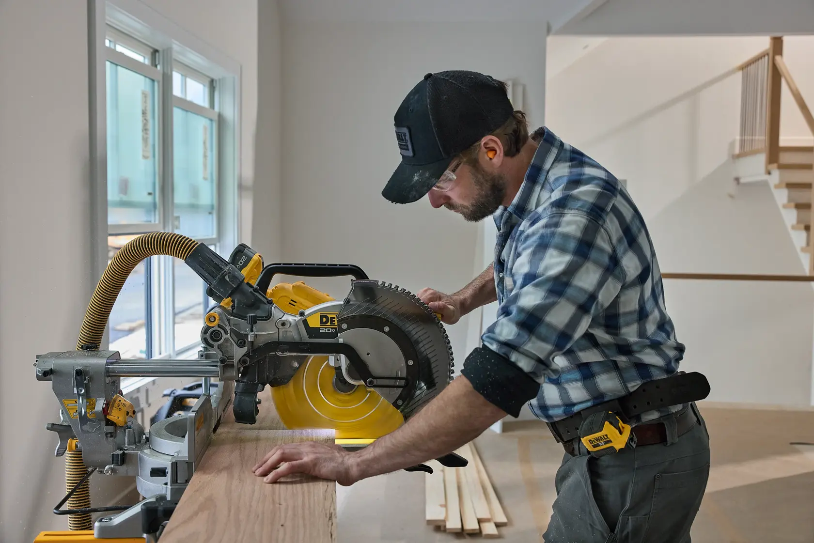 A person using a DEWALT miter saw to cut a piece of wood indoors. The individual is wearing a plaid shirt, a black cap, and a tool belt. The saw is connected to a yellow vacuum hose for dust collection. There are windows and a staircase visible in the background.