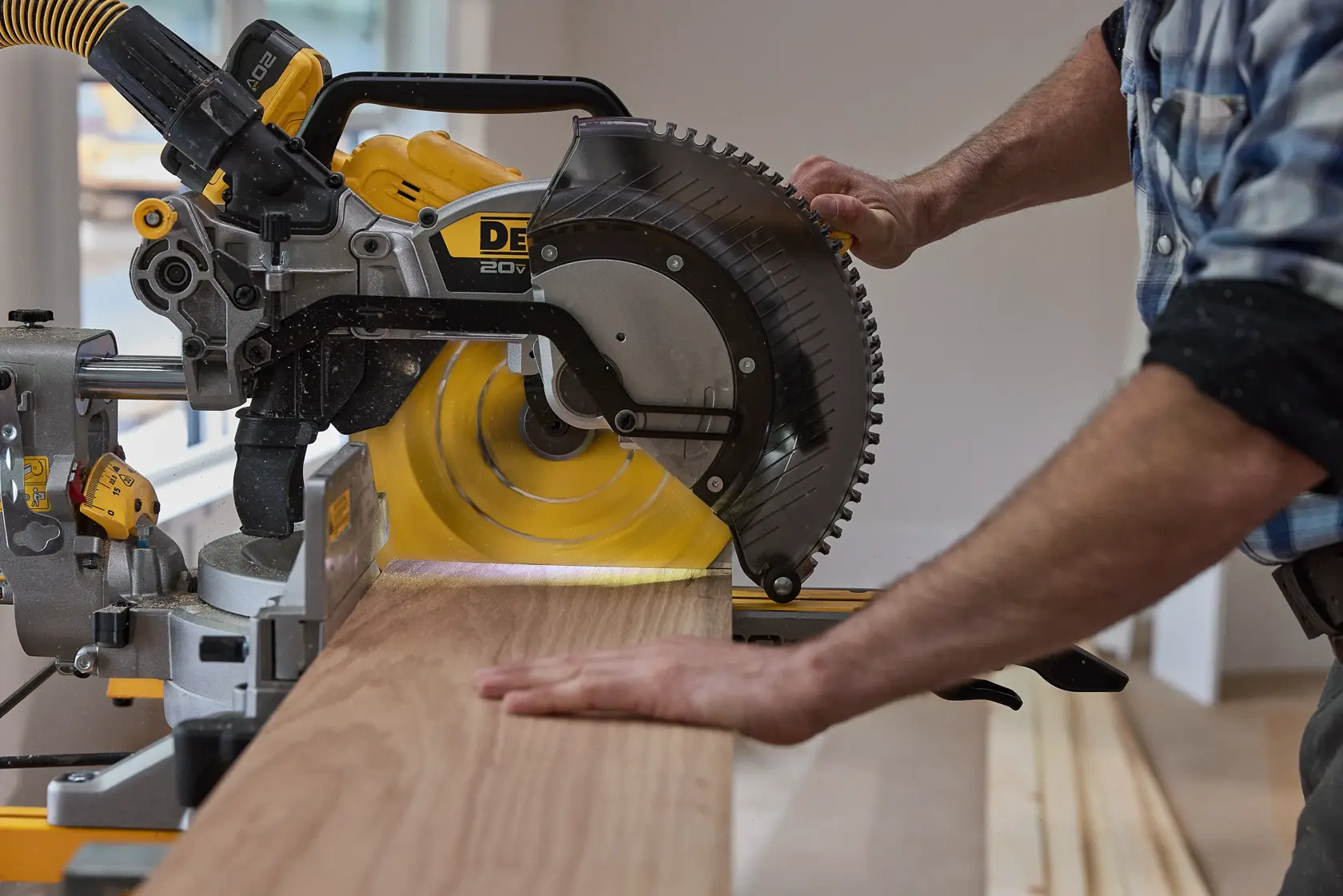 A person cutting a wooden plank using a DEWALT miter saw inside a workshop.