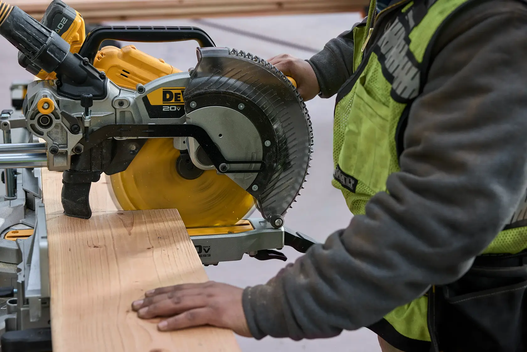 A person using a DEWALT power miter saw to cut a piece of wood. The person is wearing a safety vest and operating the saw in a workshop setting.