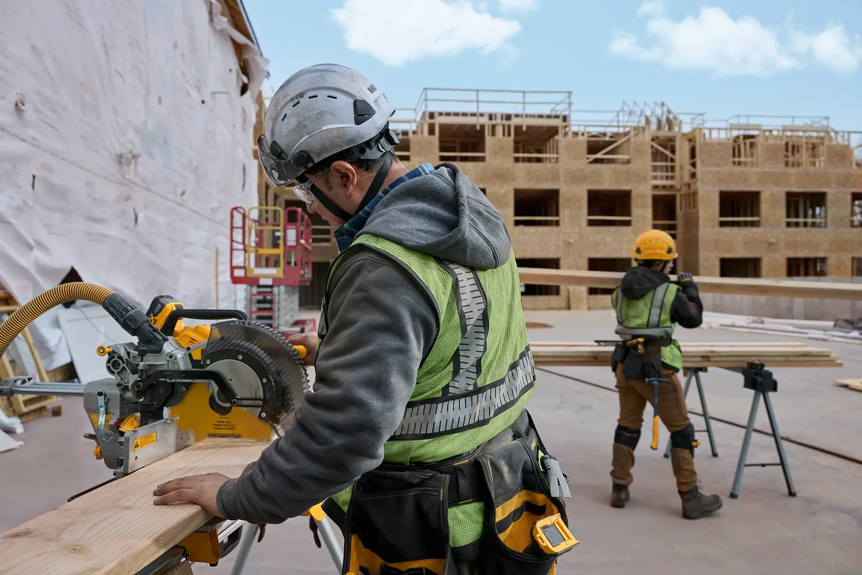 Two construction workers at an outdoor building site. One worker in the foreground is using a DEWALT miter saw to cut a wooden board, wearing a safety vest and helmet. Another worker in the background is handling wood planks, with a partially built wooden structure visible.