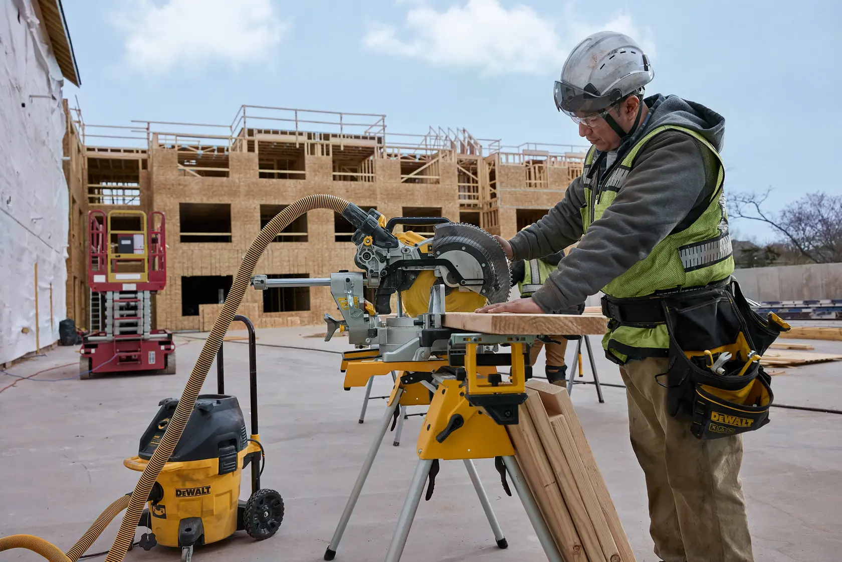 A construction worker wearing safety gear operates a DEWALT miter saw on a stand at a building site. Wooden framework of a multi-story building is visible in the background, along with other construction equipment and tools.