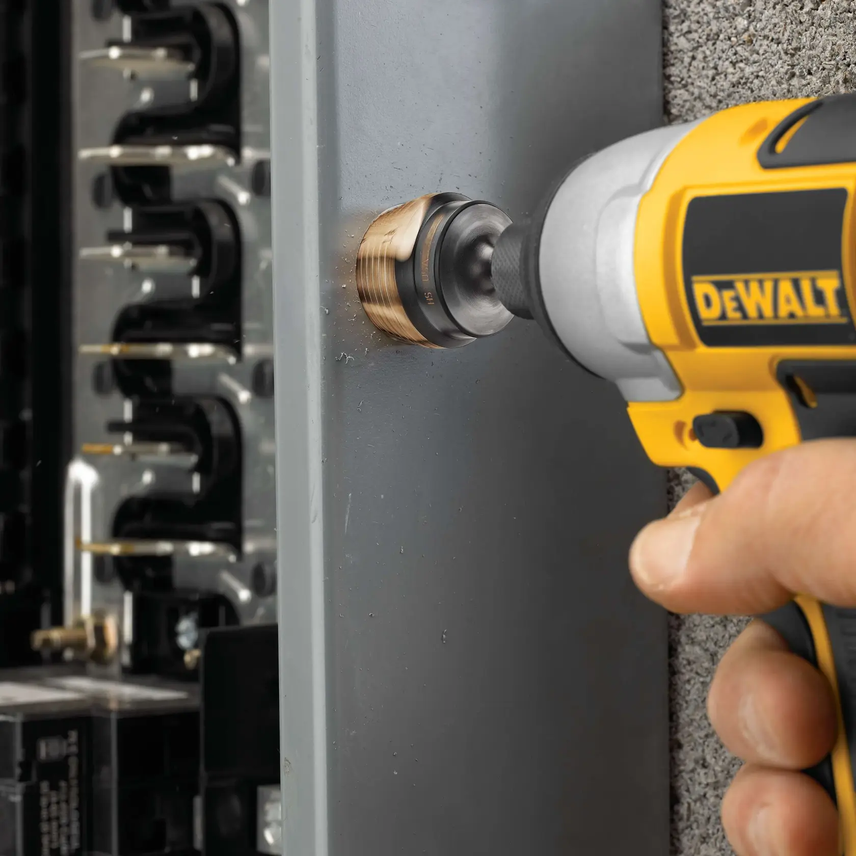 Close-up of a DEWALT power drill with a hole saw attachment being used to drill into a gray metal electrical panel.