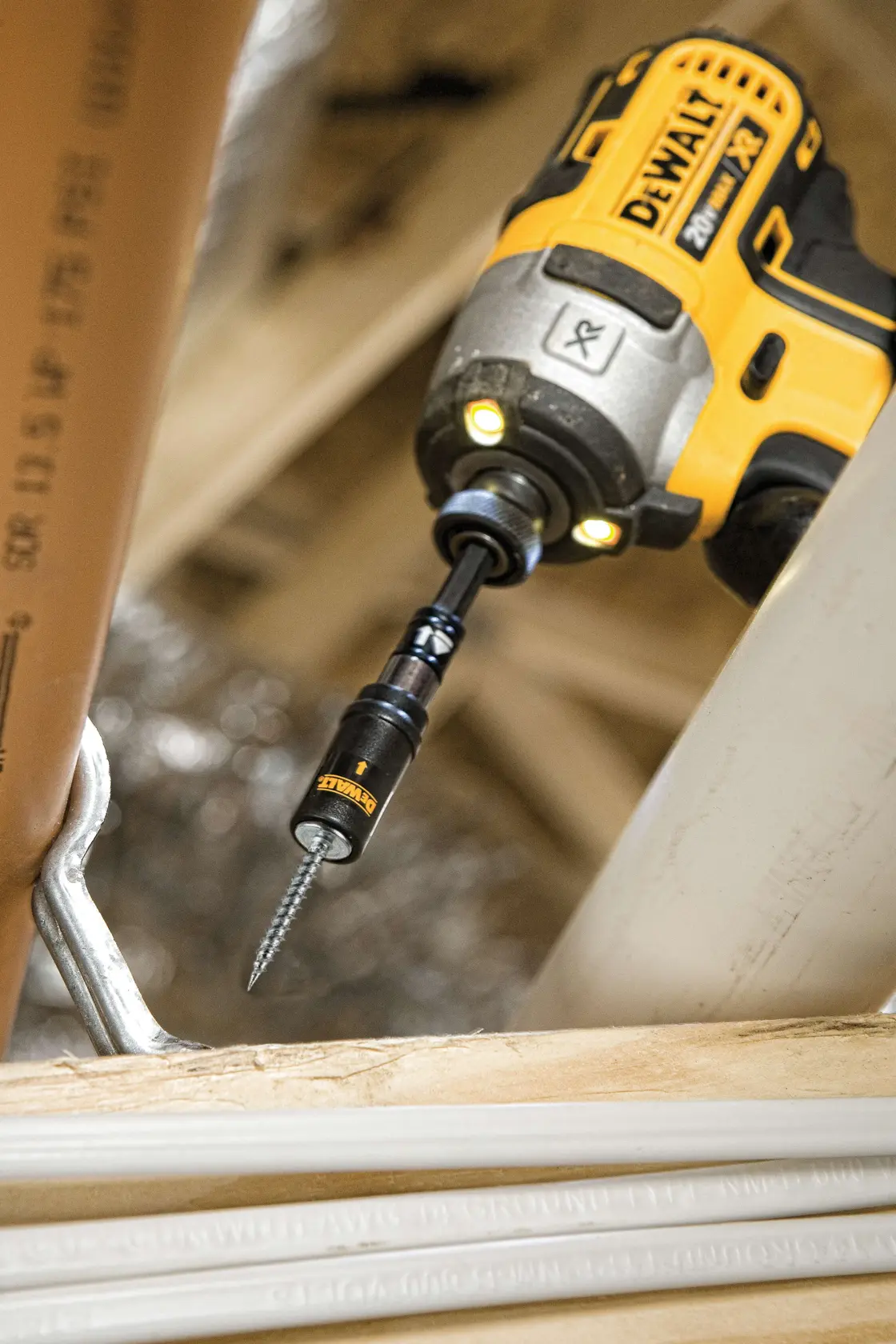 Une perceuse électrique DEWALT avec un embout pour vis est utilisée pour insérer une vis dans une surface en bois dans un environnement de construction. La perceuse est jaune et noire, avec le logo DEWALT visible.