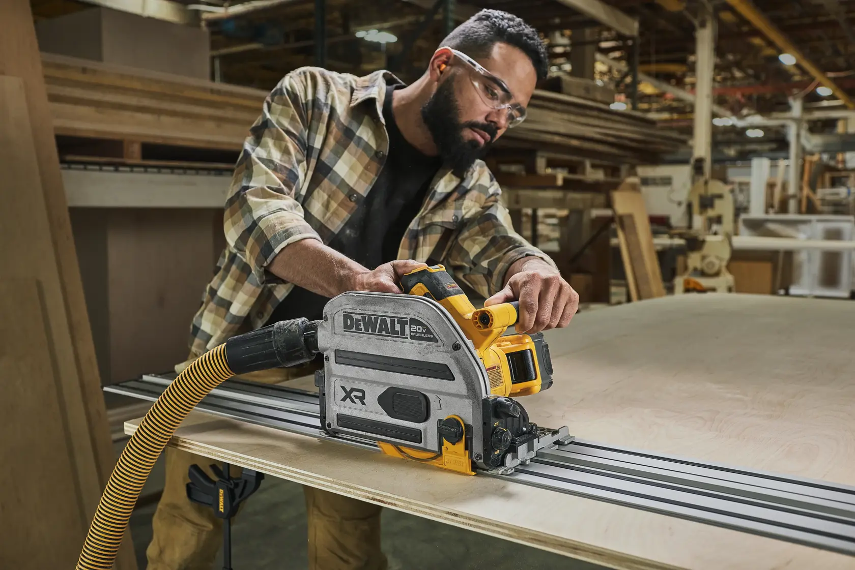 Une personne utilise une scie à onglet sans fil DEWALT, modèle DWA261248TS, pour couper une grande planche de bois sur une table d'atelier. La scie est fixée à un rail de guidage et reliée à un tuyau d'extraction de poussière.